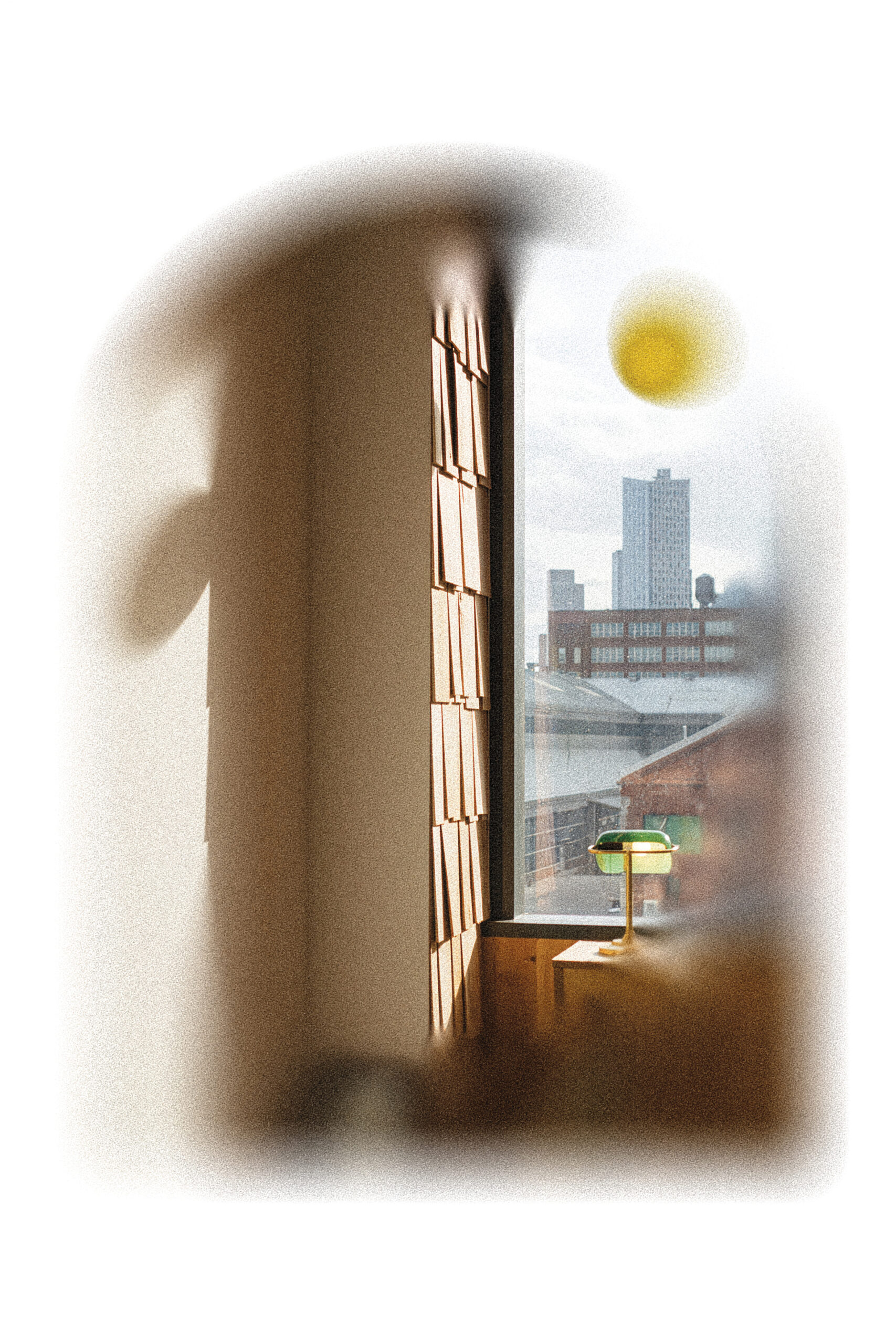 A soft-focus image of an interior space featuring a window with a view of city buildings. Sunlight casts a warm glow on the walls and a small table lamp sits on a light-colored surface nearby. The wall beside the window is adorned with textured wooden panels. The atmosphere is calm and inviting.