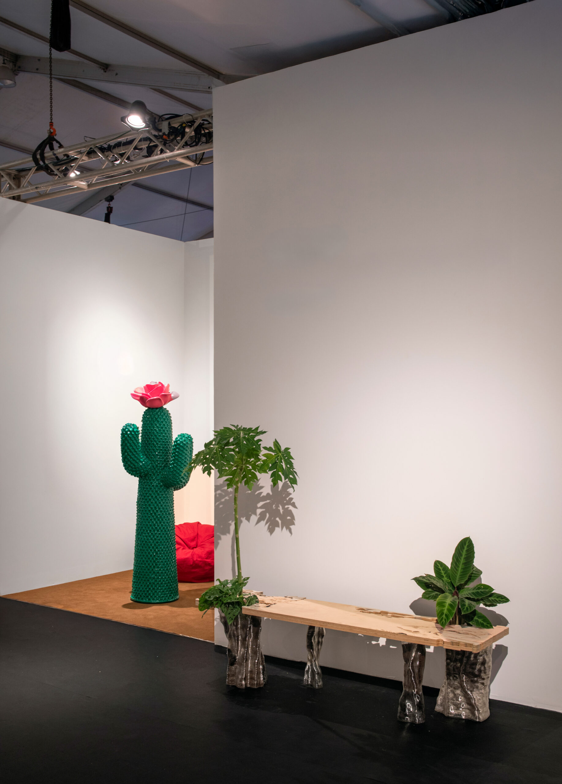 In an art fair booth, a large green cactus sculpture with a flower on top. To the right, there is a wooden bench with two planters on either end, one containing a leafy plant and the other featuring a small plant. A red bean bag is visible in the background, against a white wall.