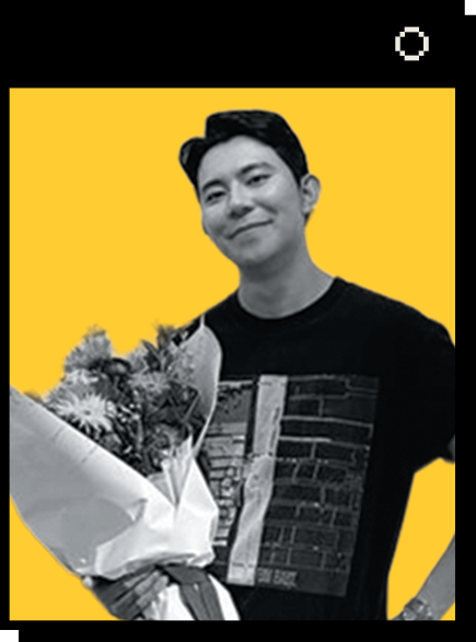 Portrait of Suhyeon Lee against a yellow background. Suhyeon smiles, holding a large bouquet of flowers wrapped in white paper. He wears a black T-shirt with a design featuring bricks and abstract shapes.