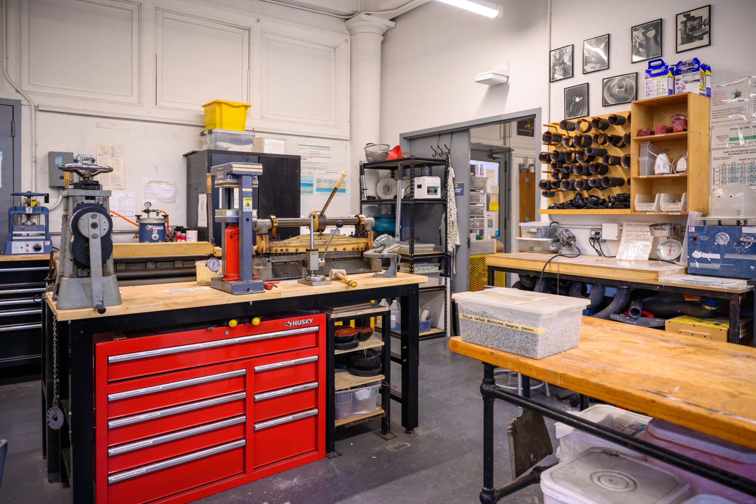 A workshop interior featuring various tools and equipment. Prominent in the center is a workbench with a red toolbox and several machines, including a press and a rolling mill. Shelves on the walls are lined with tools, jars, and tubes. A bulletin board displays paperwork, and photographs are framed on the wall. The floor is gray, and the space appears organized and functional for hands-on projects.