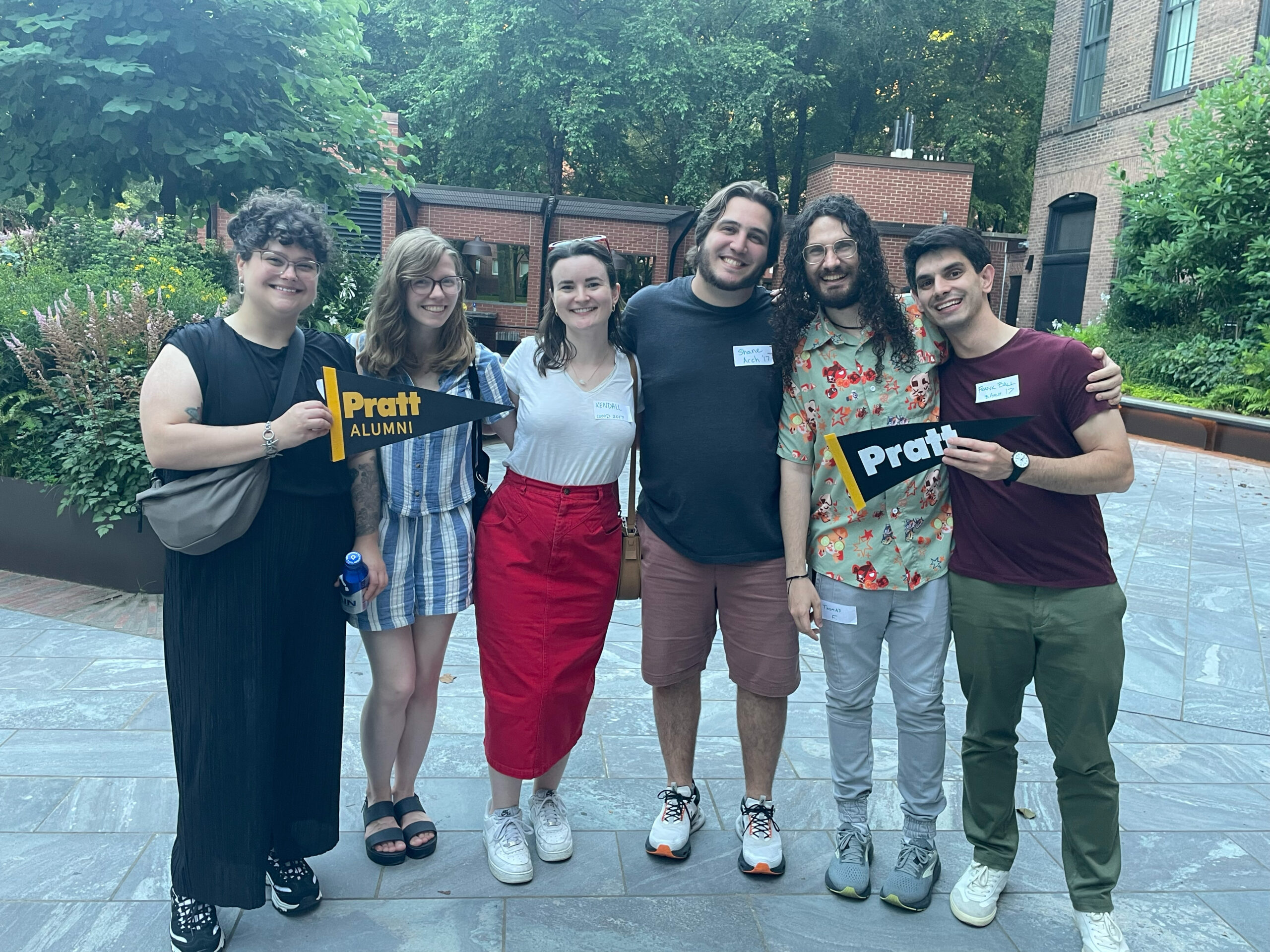 A group of six people pose together outdoors, smiling and holding a black and yellow "Pratt ALUMNI" pennant. The setting includes greenery and brick buildings in the background. The individuals are dressed casually, and some have name tags on their shirts. The atmosphere appears friendly and celebratory.