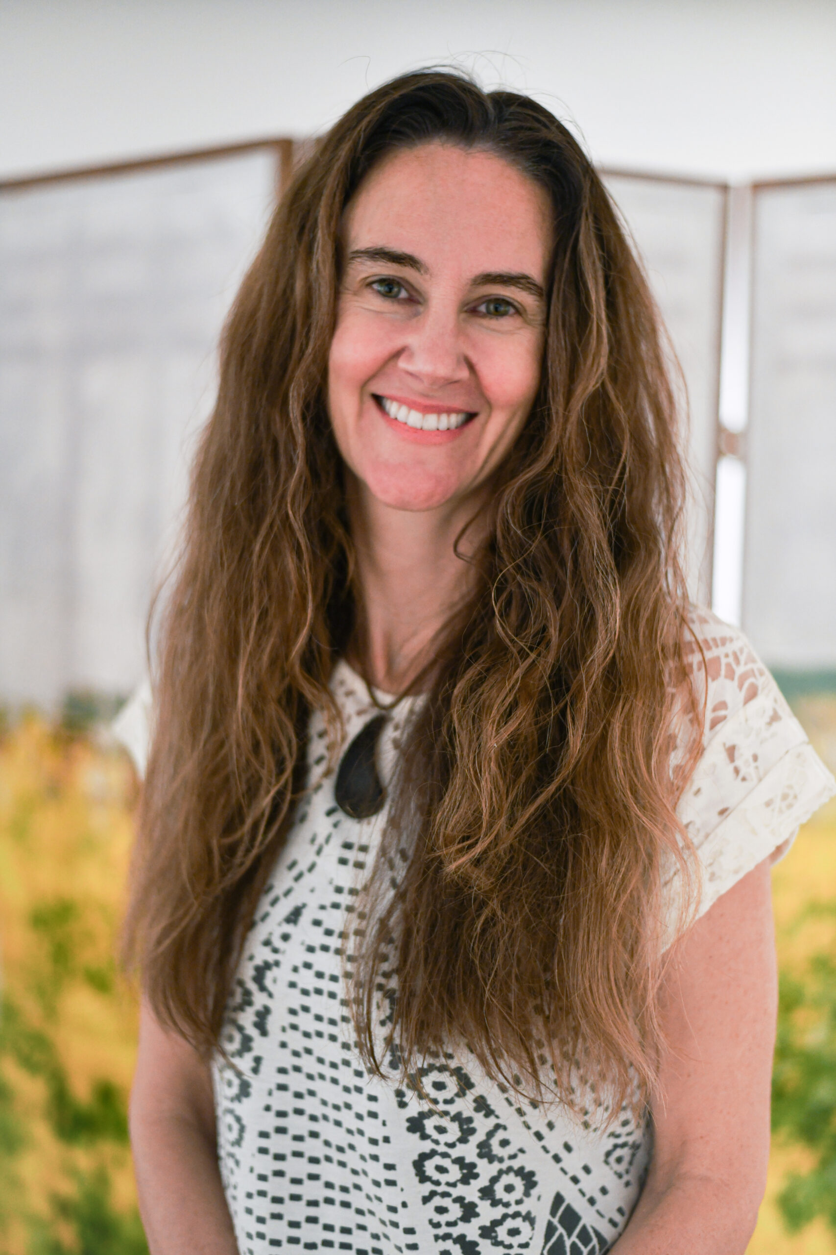 A woman with long, wavy dark hair smiles at the camera. She is wearing a white top with a decorative black pattern. In the background, there are blurred images of greenery and a wooden partition, suggesting an indoor setting.