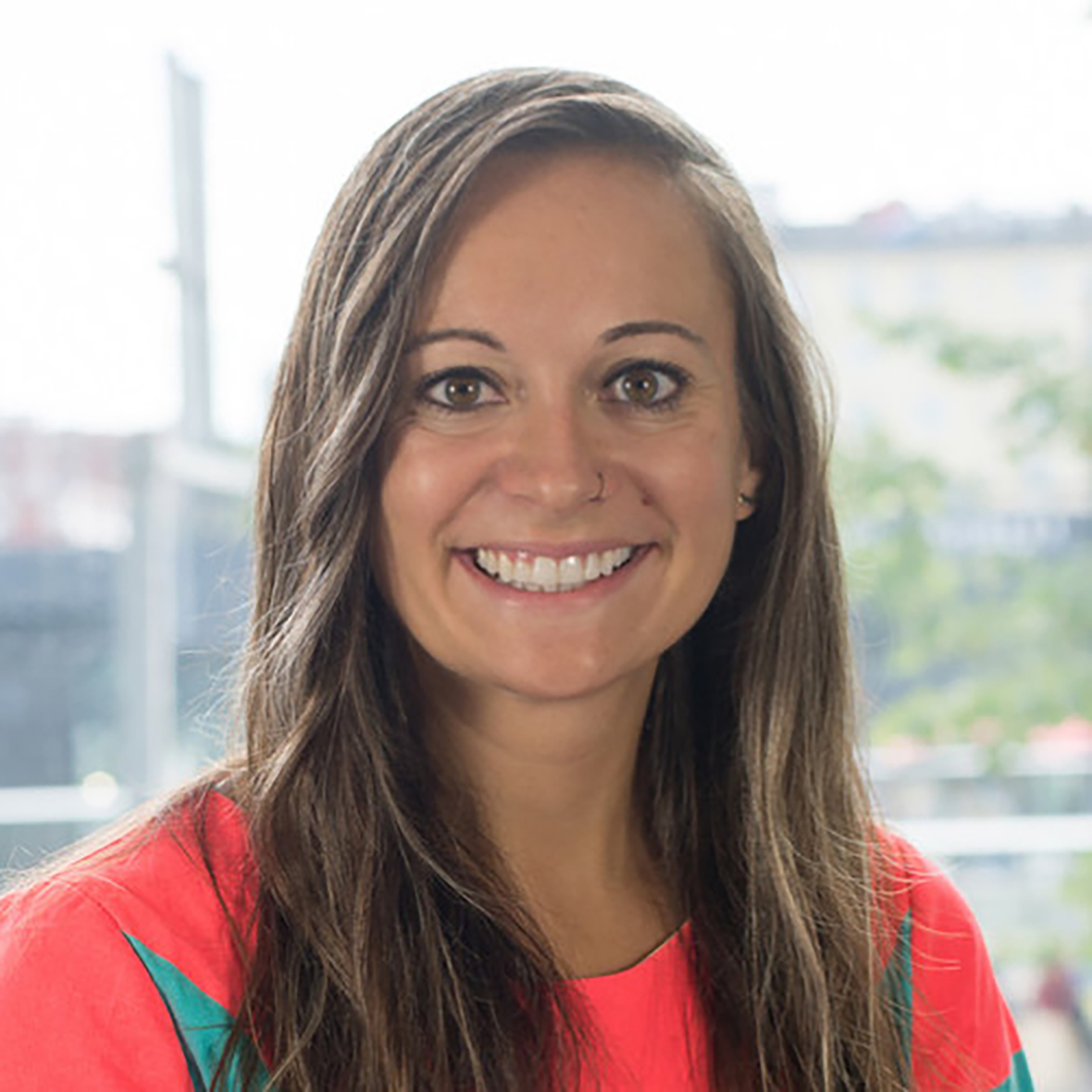 A smiling woman with long, wavy brown hair is wearing a bright coral shirt. She has a nose ring and is in front of a bright, open window with greenery visible in the background. The overall setting appears light and cheerful.