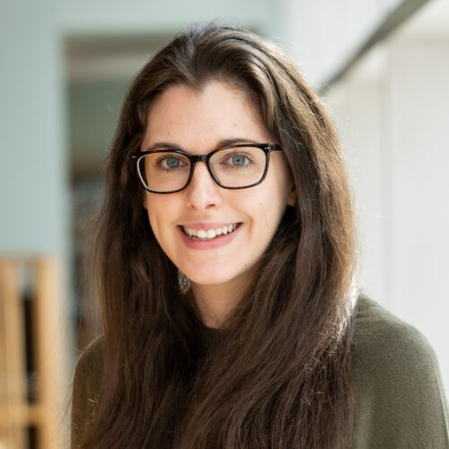 A young woman with long, dark hair and glasses smiles at the camera. She is wearing a green sweater and is positioned in a well-lit indoor space. Natural light highlights her features, and there are blurred wooden structures in the background.