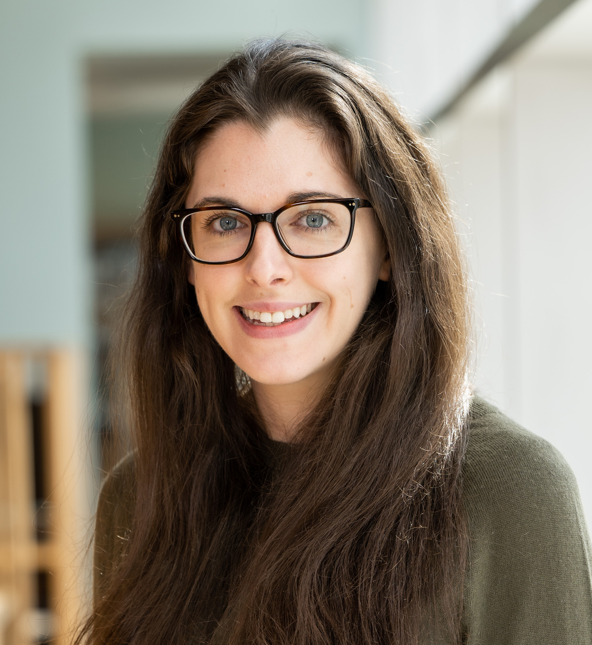 A young woman with long, dark hair and glasses smiles at the camera. She is wearing a green sweater and is positioned in a well-lit indoor space. Natural light highlights her features, and there are blurred wooden structures in the background.