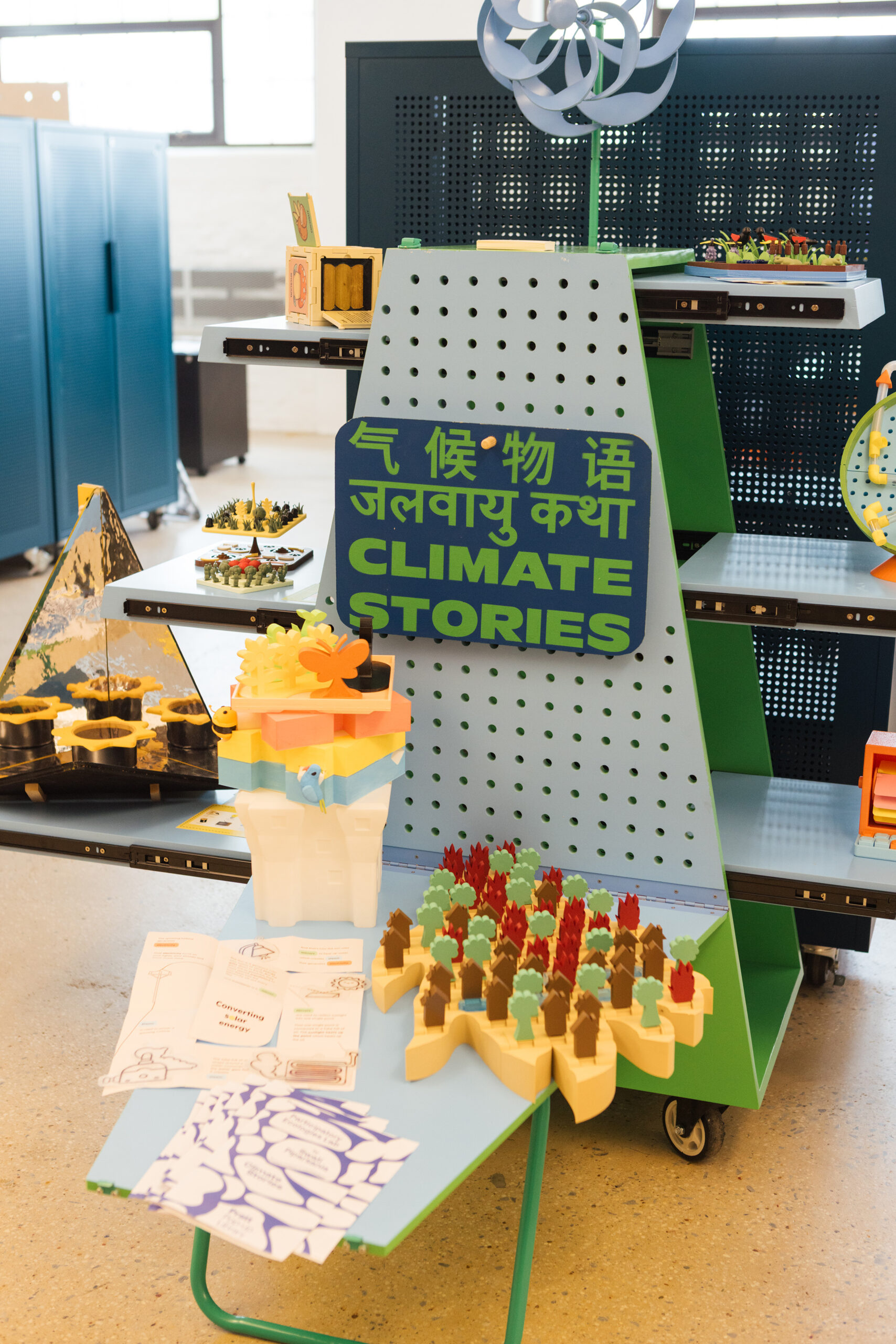 A colorful display showing models related to climate themes, titled "CLIMATE STORIES." The exhibit features various structures made from plastic, including a layered building, a pyramid-like design with a plant motif, and a landscape with trees and flames. Educational materials such as pamphlets and prints are displayed on the table, and there are colorful elements like bees and birds incorporated into the models. The background includes storage cabinets, giving it a vibrant educational atmosphere.