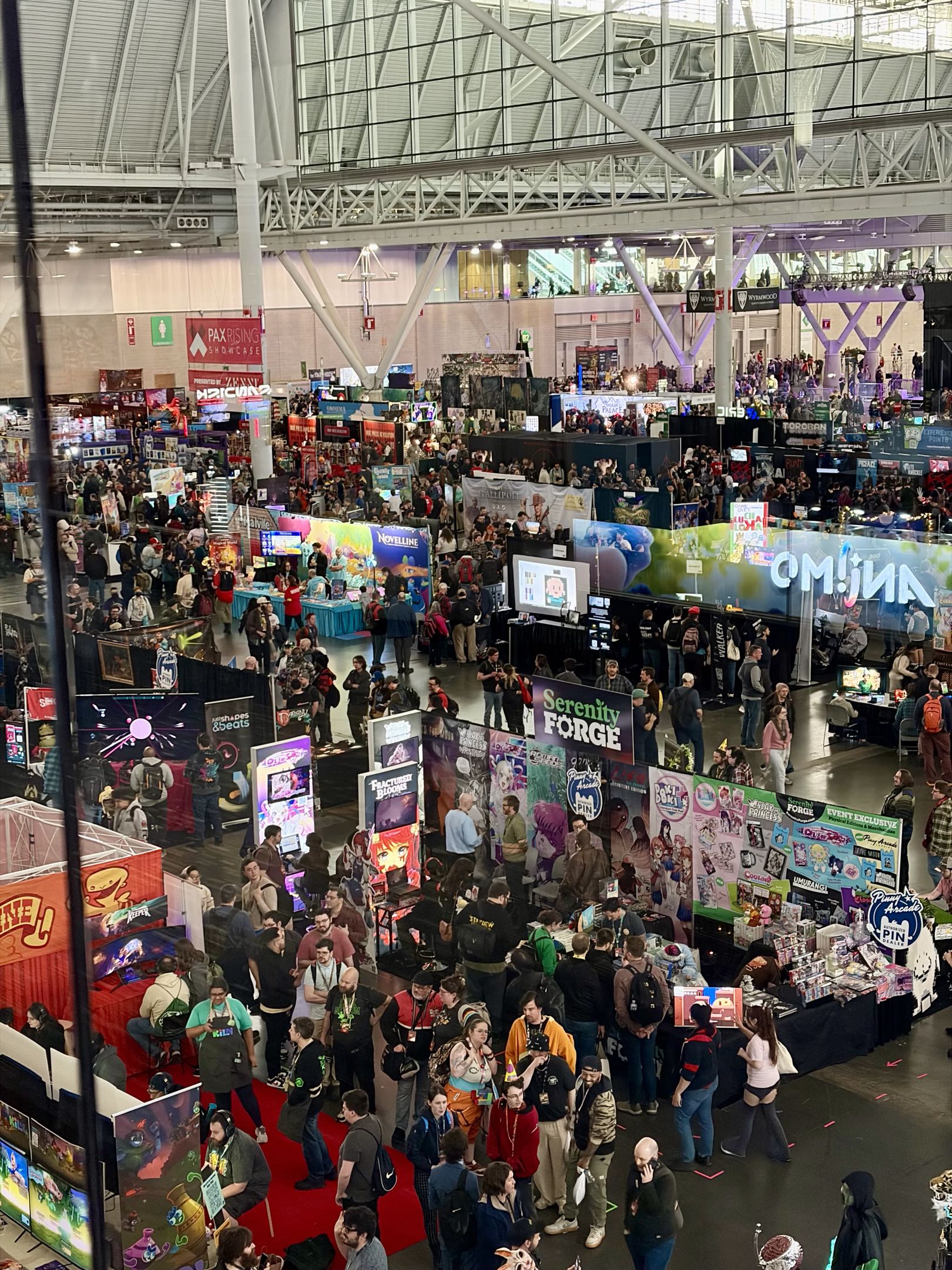 A bustling convention hall filled with booths and displays. Attendees browse various vendors showcasing games and technology. Colorful banners highlight different exhibits. The crowd includes people of diverse ages and backgrounds, with some wearing gaming-related apparel. The venue's modern architecture is visible overhead.