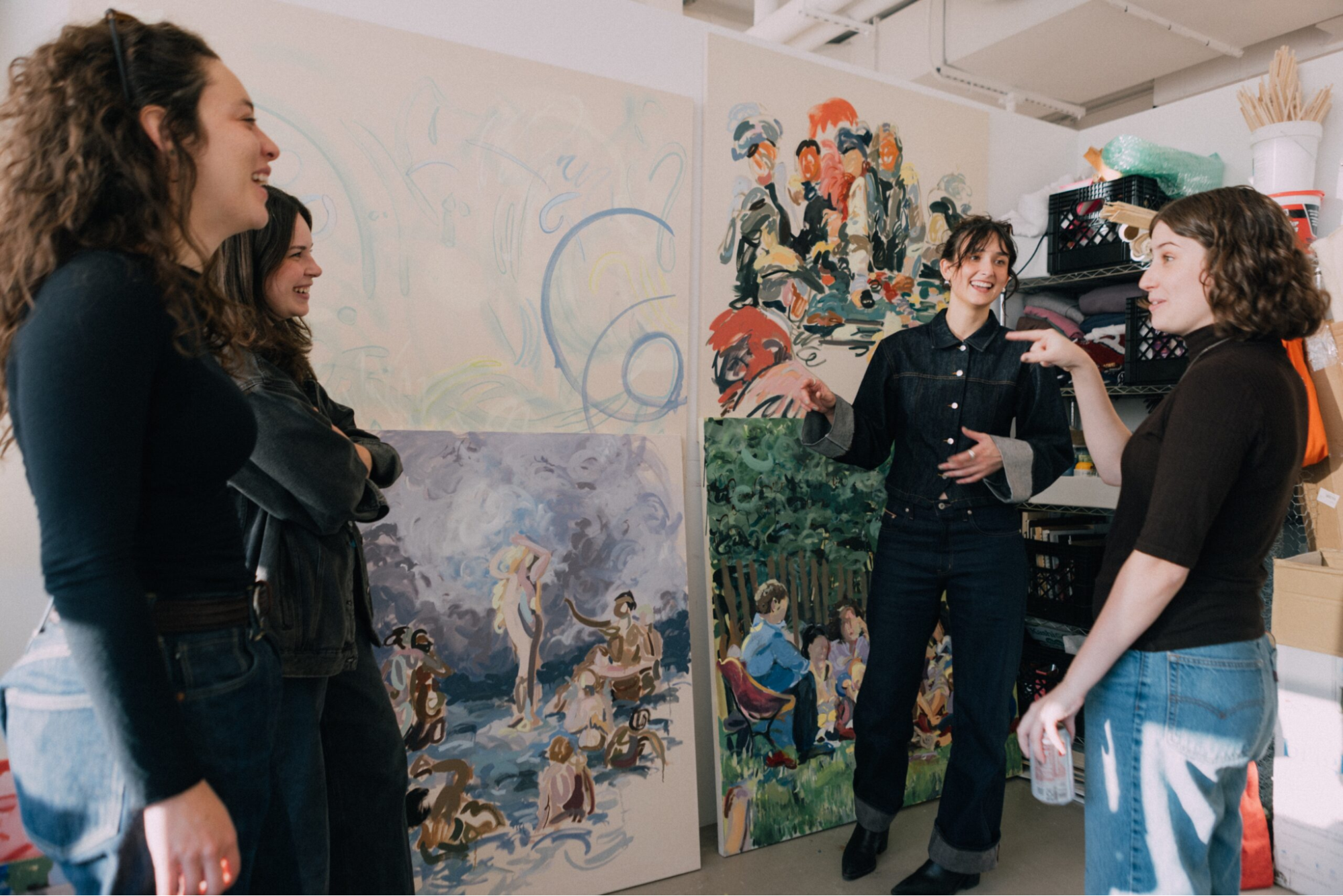 Four women are engaged in conversation in an art studio filled with large colorful paintings on the walls. They are smiling and interacting animatedly. The background features various artworks, including abstract designs and scenes with figures, while art supplies and materials are visible in the cluttered space.