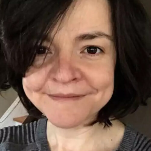 A person with short, dark hair is smiling gently at the camera. They are wearing a gray sweater with ribbed texture and button details on one shoulder. The background is blurred, showing a hint of a staircase and room decor.