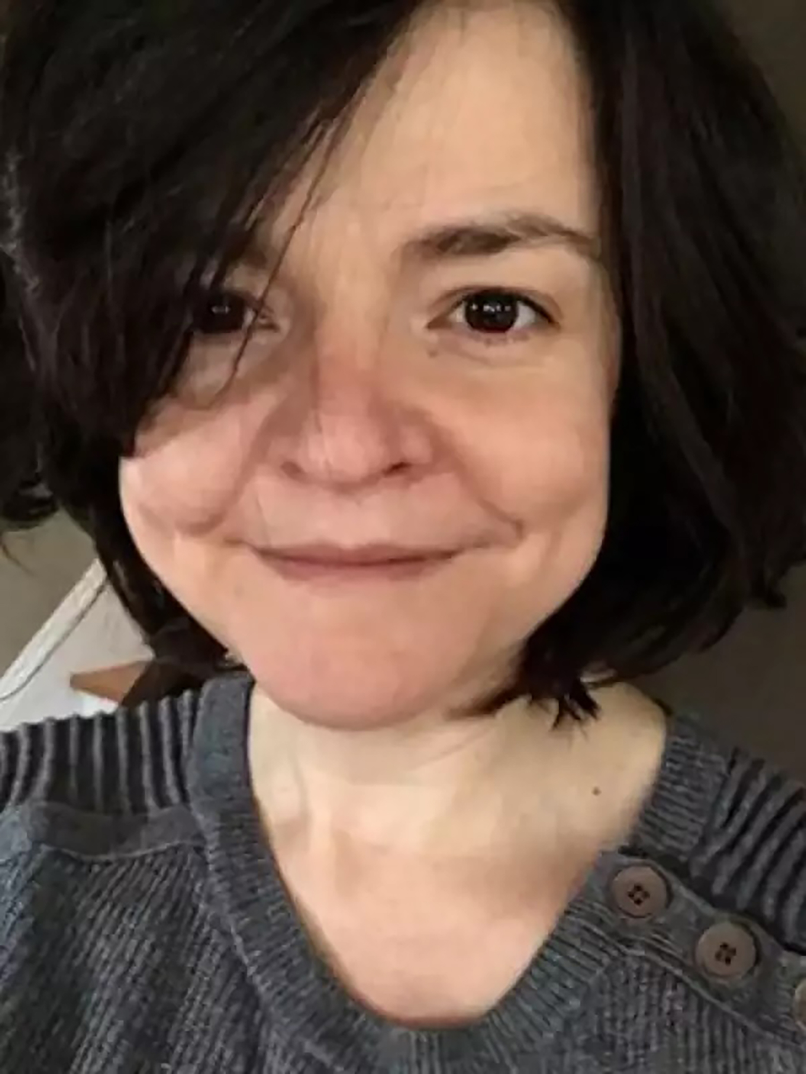 A person with short, dark hair is smiling gently at the camera. They are wearing a gray sweater with ribbed texture and button details on one shoulder. The background is blurred, showing a hint of a staircase and room decor.