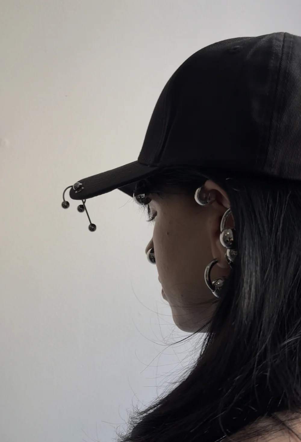 A model, seen from over her shoulder, wears a black baseball cap. The brim is pierced with silver industrial hardware.