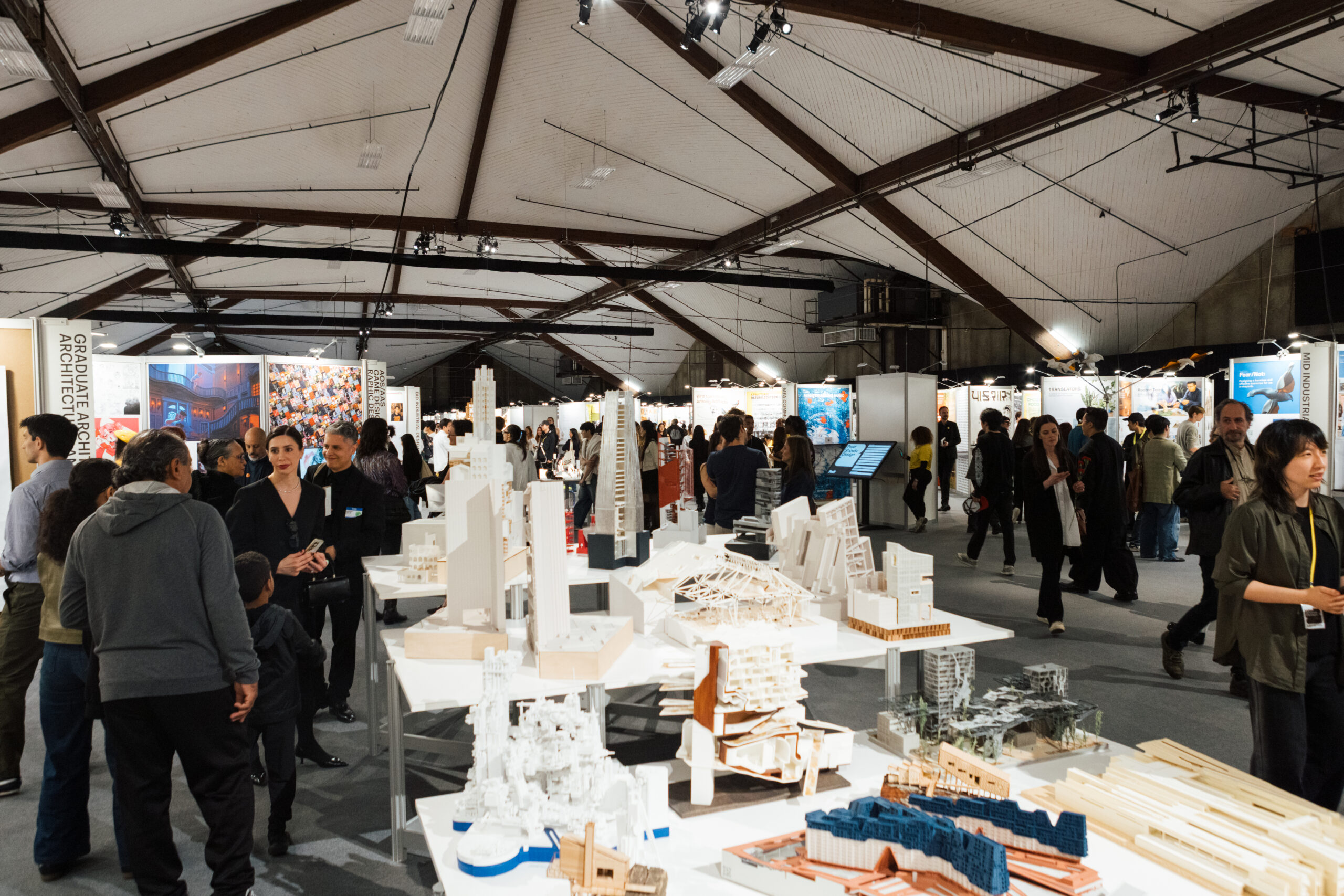 A large indoor exhibition space filled with people engaging with architectural models and displays. The area features several stands showcasing architectural work, with models on tables in the foreground. The ceiling has wooden beams, and the ambiance appears busy and collaborative, highlighting the architectural community.
