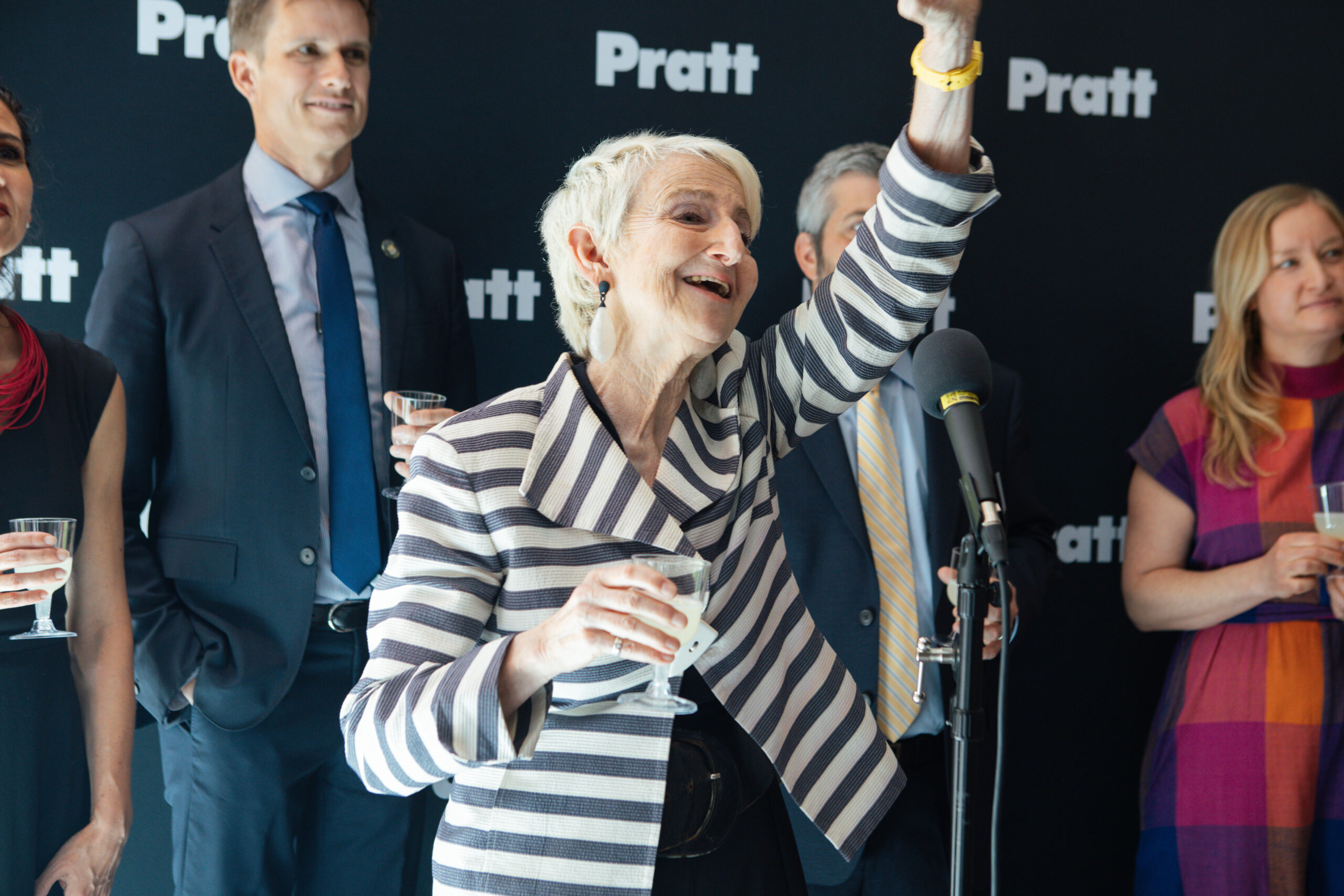 A woman with short grey hair and a striped blazer holds a glass while raising her hand in a celebratory gesture. She is smiling and surrounded by several people in formal attire, standing in front of a backdrop that reads "Pratt." The event appears to be festive or commemorative in nature.
