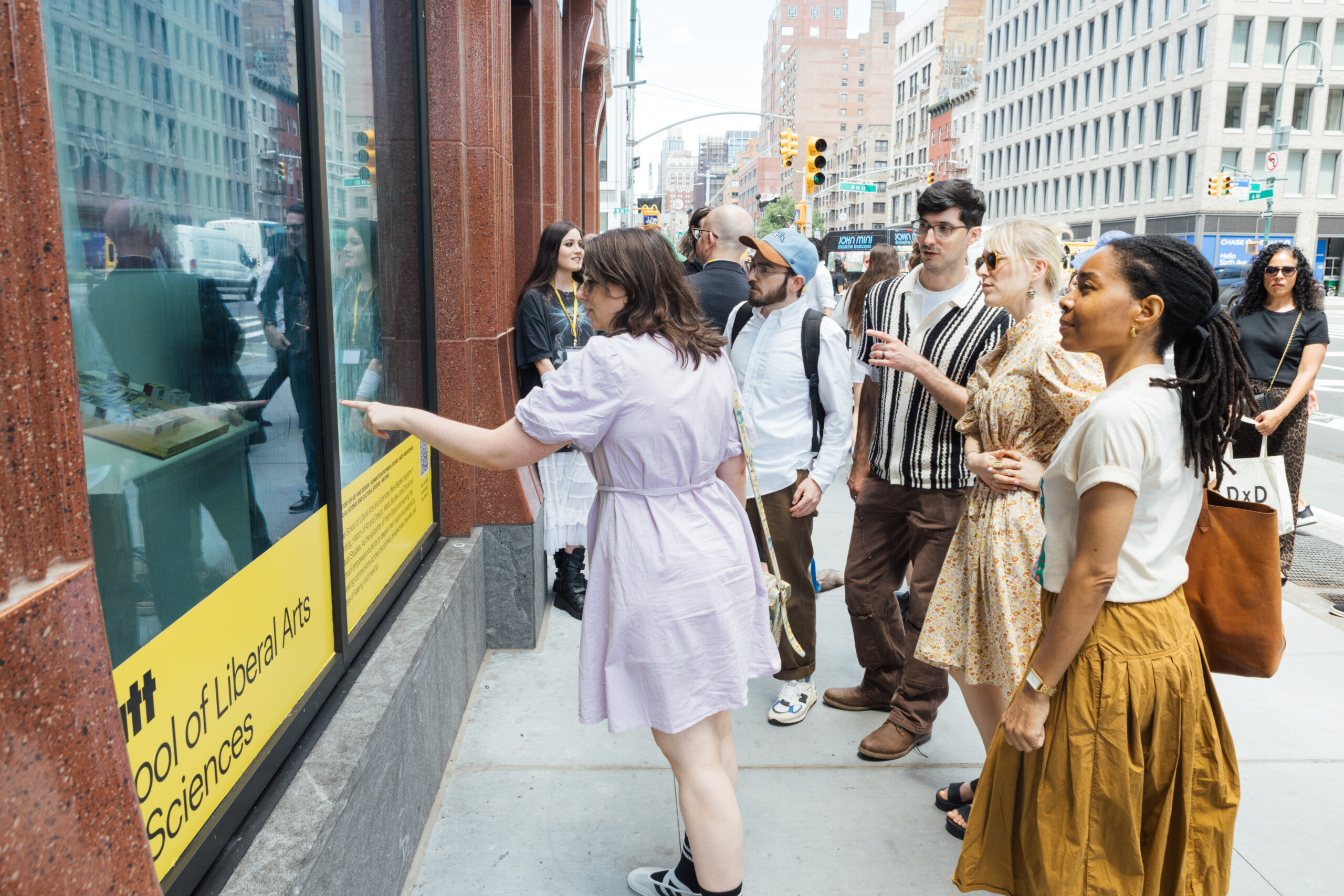 A small group of people stands on a city sidewalk, looking at a display in a storefront window. One person, wearing a lavender dress, points towards the display while others watch attentively. The crowd consists of individuals in various casual outfits, with some holding drinks. The storefront features a yellow sign with text indicating an affiliation with a School of Liberal Arts and Sciences.