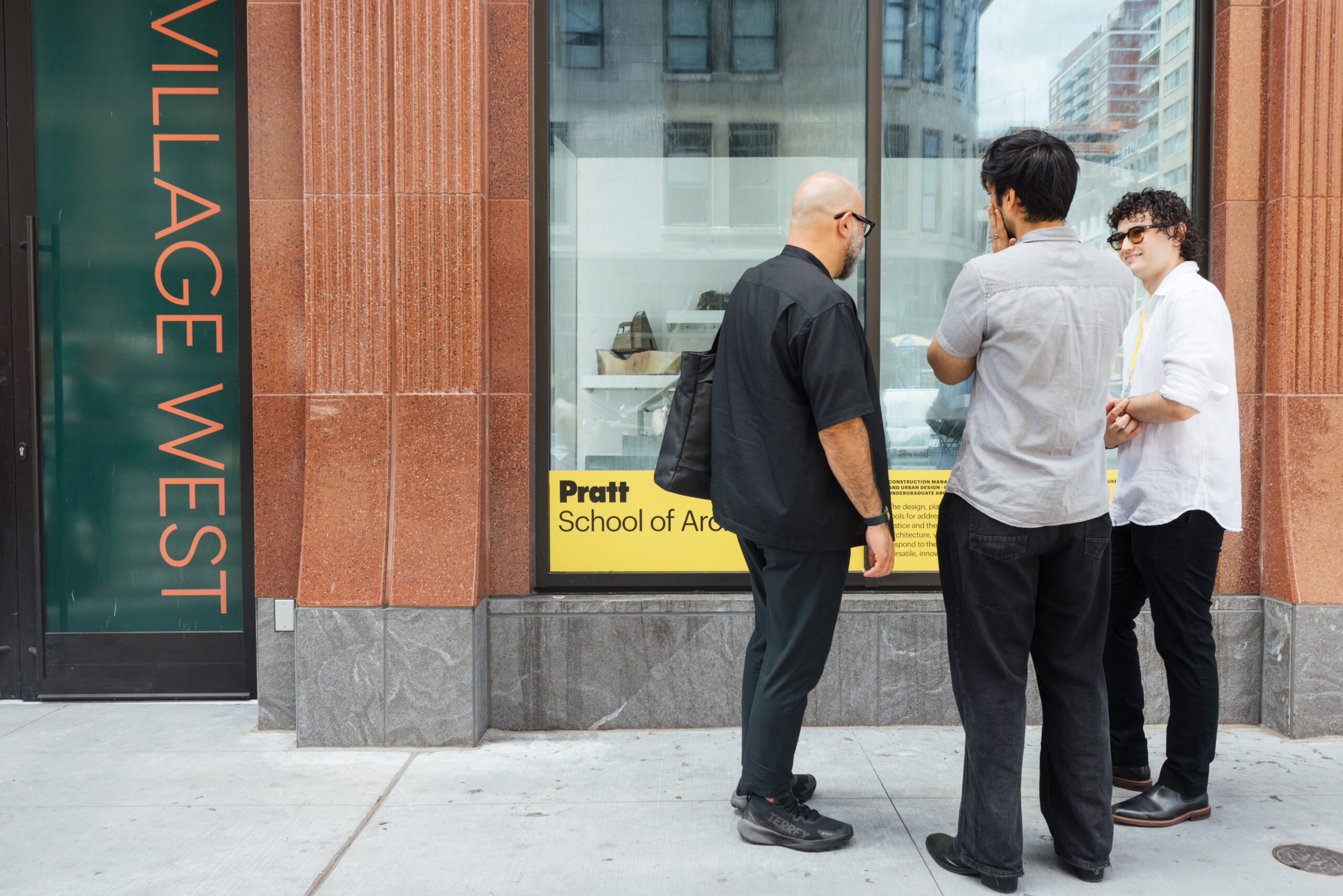 Three men stand outside a building with a "VILLAGE WEST" sign. They are engaged in conversation, looking at a window display featuring the "Pratt School of Architecture" with a yellow background. The men are dressed casually, with one holding a black bag. The background includes architectural features and urban scenery.