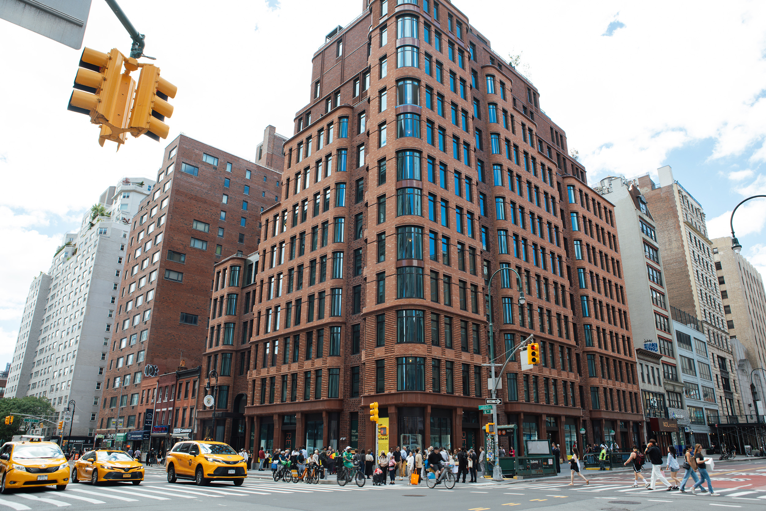 A busy urban street corner features a modern red-brick building with large windows, alongside other buildings of varying heights and styles. Yellow taxis are seen driving along the street, while pedestrians, including cyclists, cross at the intersection. Traffic lights and street signs are visible, highlighting the lively city atmosphere under a partly cloudy sky.
