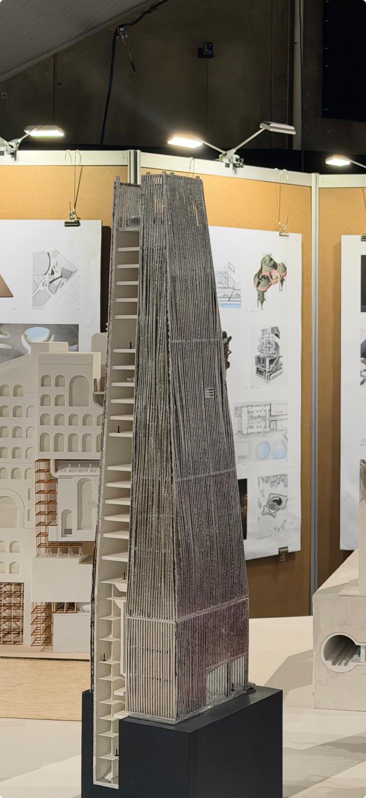 A detailed architectural model of a tall, narrow building, showcasing a textured exterior with vertical slats. The model is positioned on a black base and features visible interior floors. In the background, there are multiple architectural drawings and sketches displayed on a panel, with various structures and design concepts illustrated. The setting is well-lit, highlighting the model's intricate details.