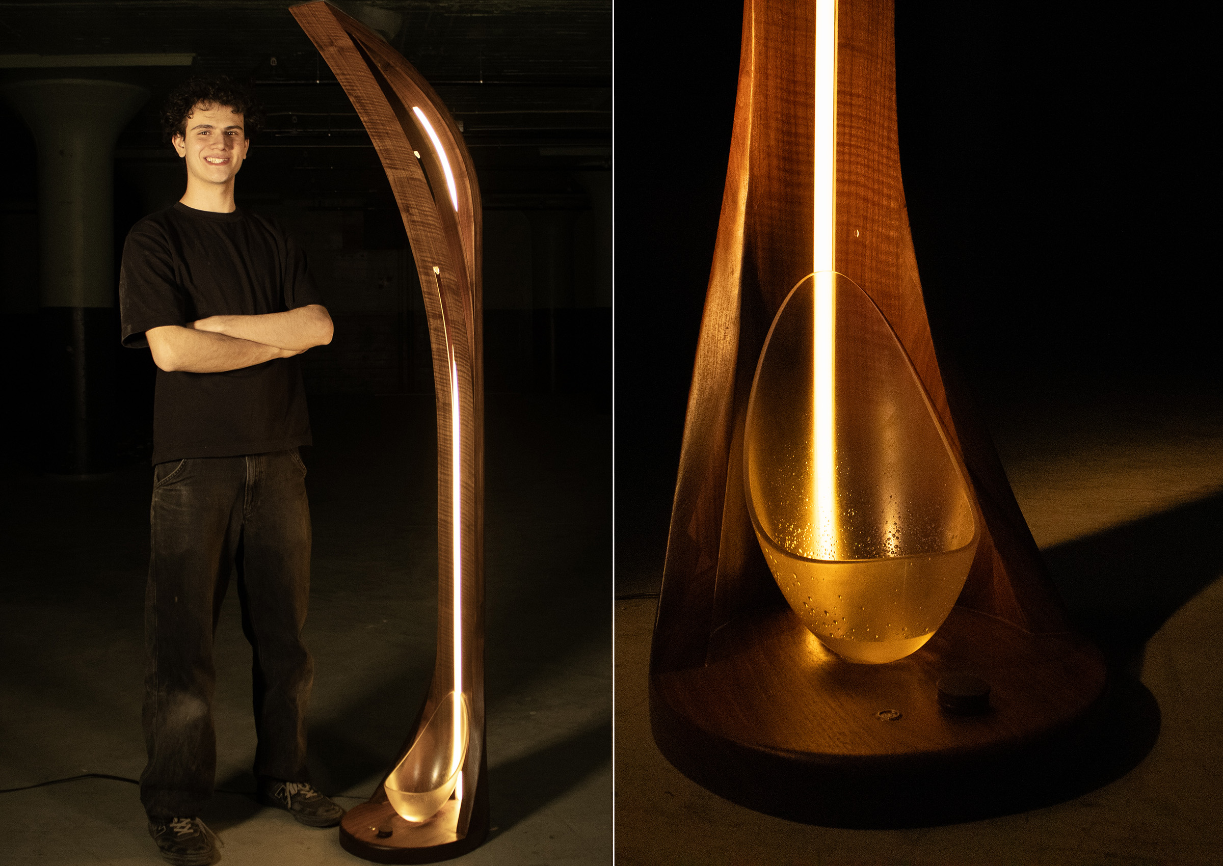 A young man stands beside a tall, curved wooden lamp that features an internal light source. The lamp is designed with a decorative, elongated shape, and includes a rounded glass element at its base, where the light shines through, creating an atmospheric glow. The setting appears dim, highlighting the lamp's design, while the man smiles confidently. The contrast of light and shadow enhances the wood textures and overall aesthetic of the lamp.