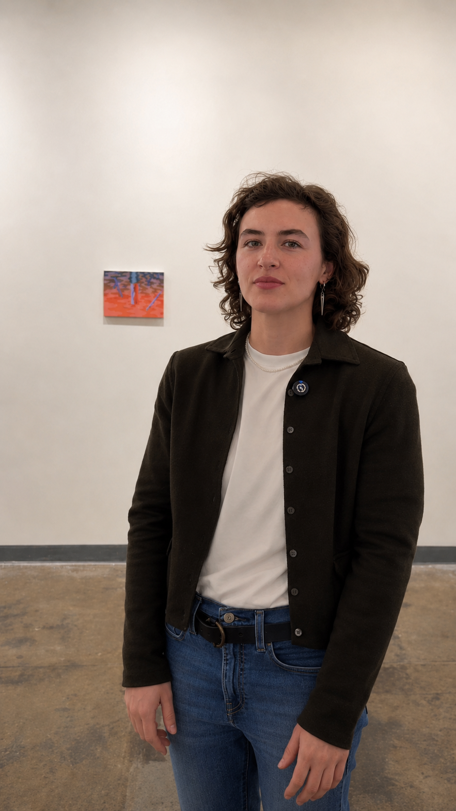 A person with short, curly hair stands in an art gallery, wearing a dark button-up jacket over a white shirt and blue jeans. They have earrings and a pin on their jacket. In the background, there's a small, colorful artwork displayed on the wall. The gallery has a simple, minimalist design with a light-colored wall and a concrete floor.