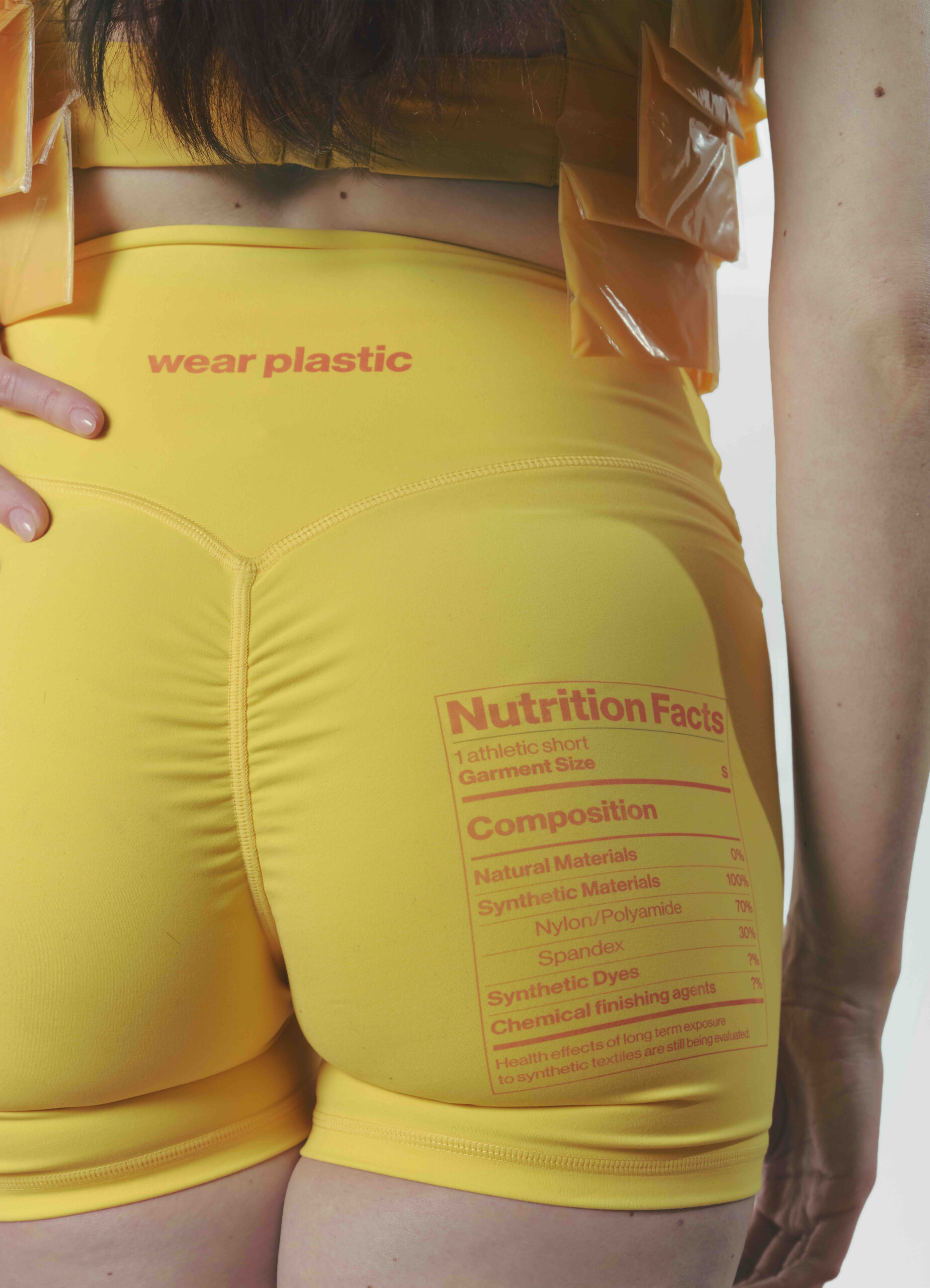 A close-up of a woman wearing bright yellow athletic shorts featuring an unconventional "Nutrition Facts" label on the back. The label lists materials, including percentages of Nylon/Polyamide and Spandex, along with a note about health effects associated with synthetic textiles. The text "wear plastic" is prominently displayed in red above the label. The model's hand rests on her hip, highlighting the garment's design.