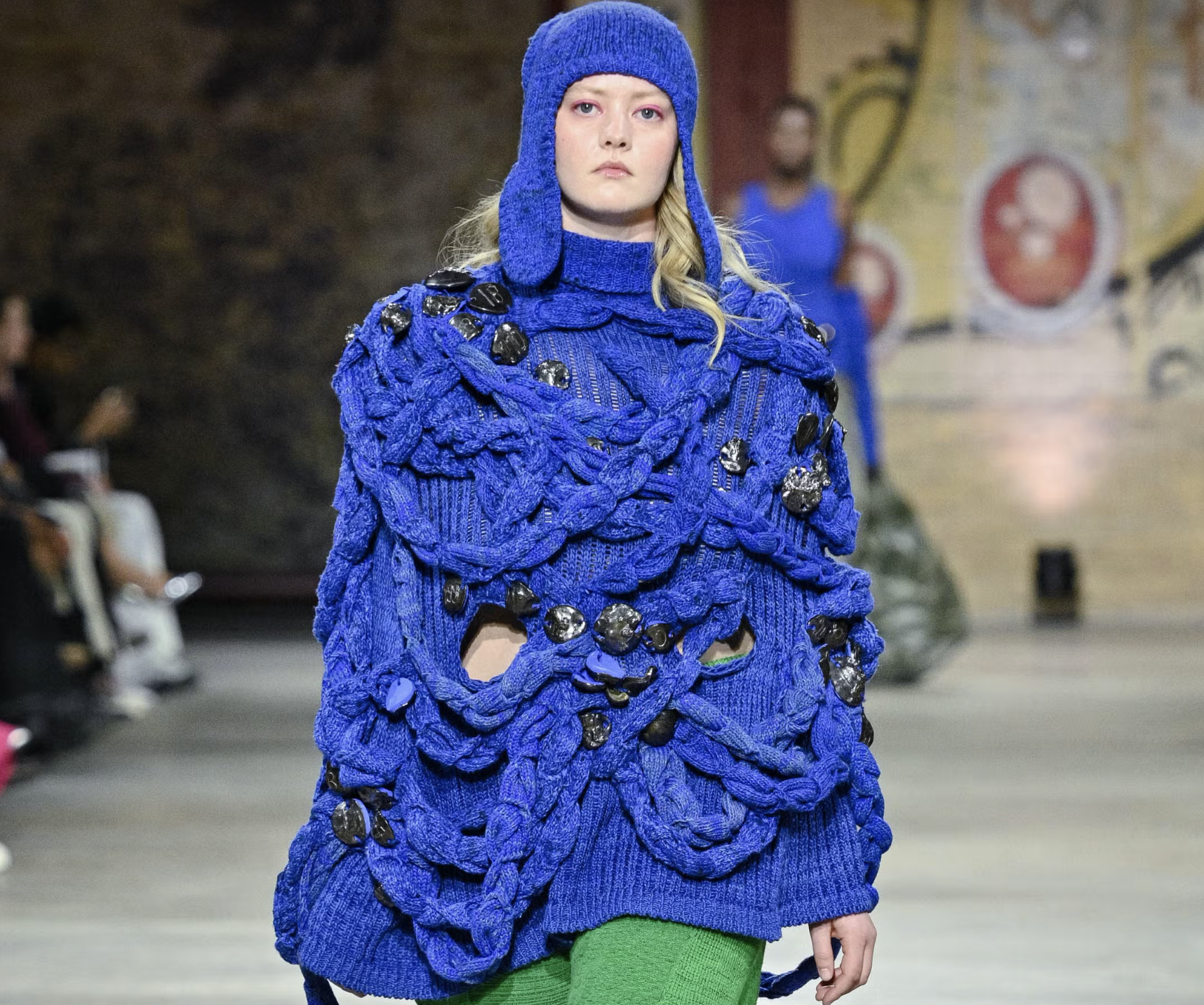 A model walks down the runway wearing an oversized, vibrant blue sweater with a textured, woven design and embellishments. The sweater covers the model's neck and features cutouts. The model pairs the sweater with bright green pants and has natural wavy hair, complemented by bold pink eye makeup. The backdrop shows a stylish venue with an audience in the background.