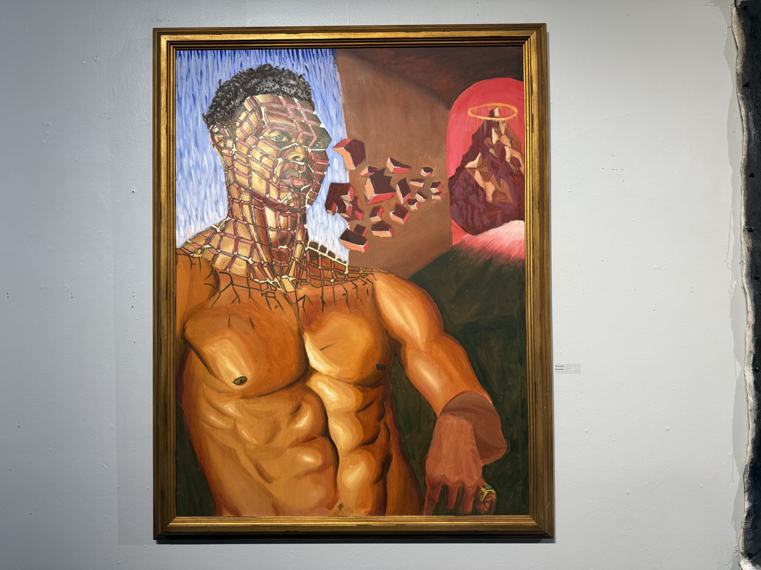 A vibrant painting features a muscular figure with a partially fragmented face, resembling a mosaic of red and brown tones. The figure's skin is a warm orange, and they have short dark hair. There are surreal elements in the background, including a red-lit area with a mountain and a halo. The artwork is framed in an ornate gold frame and displayed against a light-colored wall.