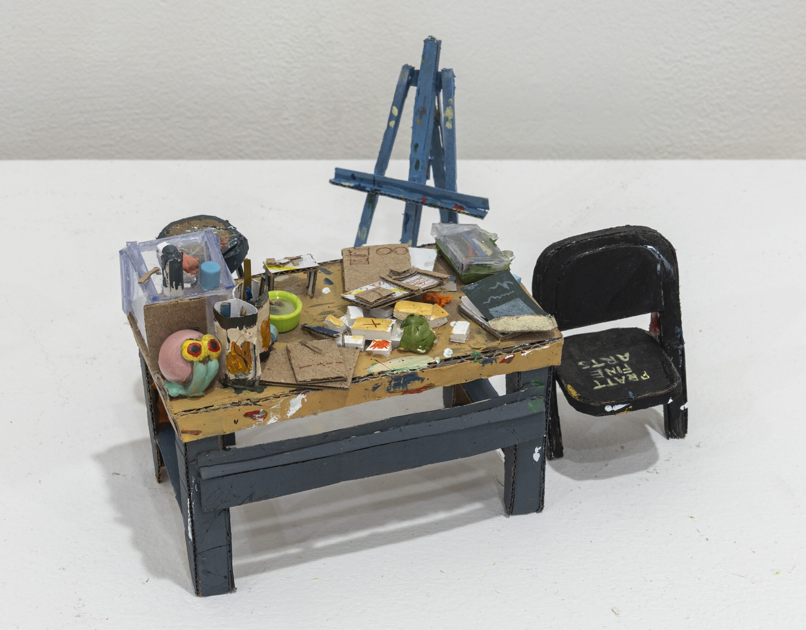 A miniature art studio featuring a cluttered table covered with various art supplies, including a transparent box, paintbrushes, paint tubes, palettes, and sketches. A colorful sculpture of a character with large glasses sits on the table. An easel is positioned at the back, and a black chair with "PAINT FINE ARTS" written on it is nearby. The scene is set against a plain white background.