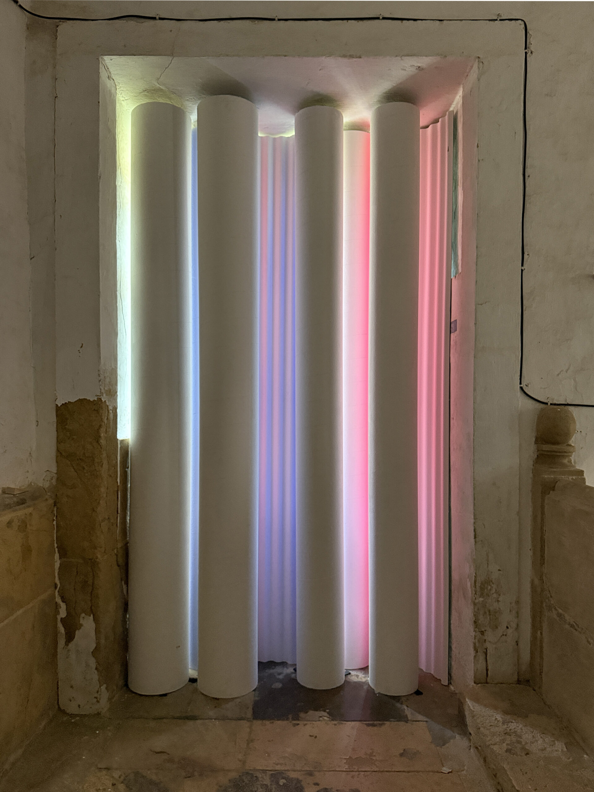 A doorway framed by six vertical, white cylindrical tubes emits soft, colorful lighting in shades of blue and pink. The space has rough, textured walls and a stone floor, emphasizing the contrast between the smooth tubes and the rustic surroundings.