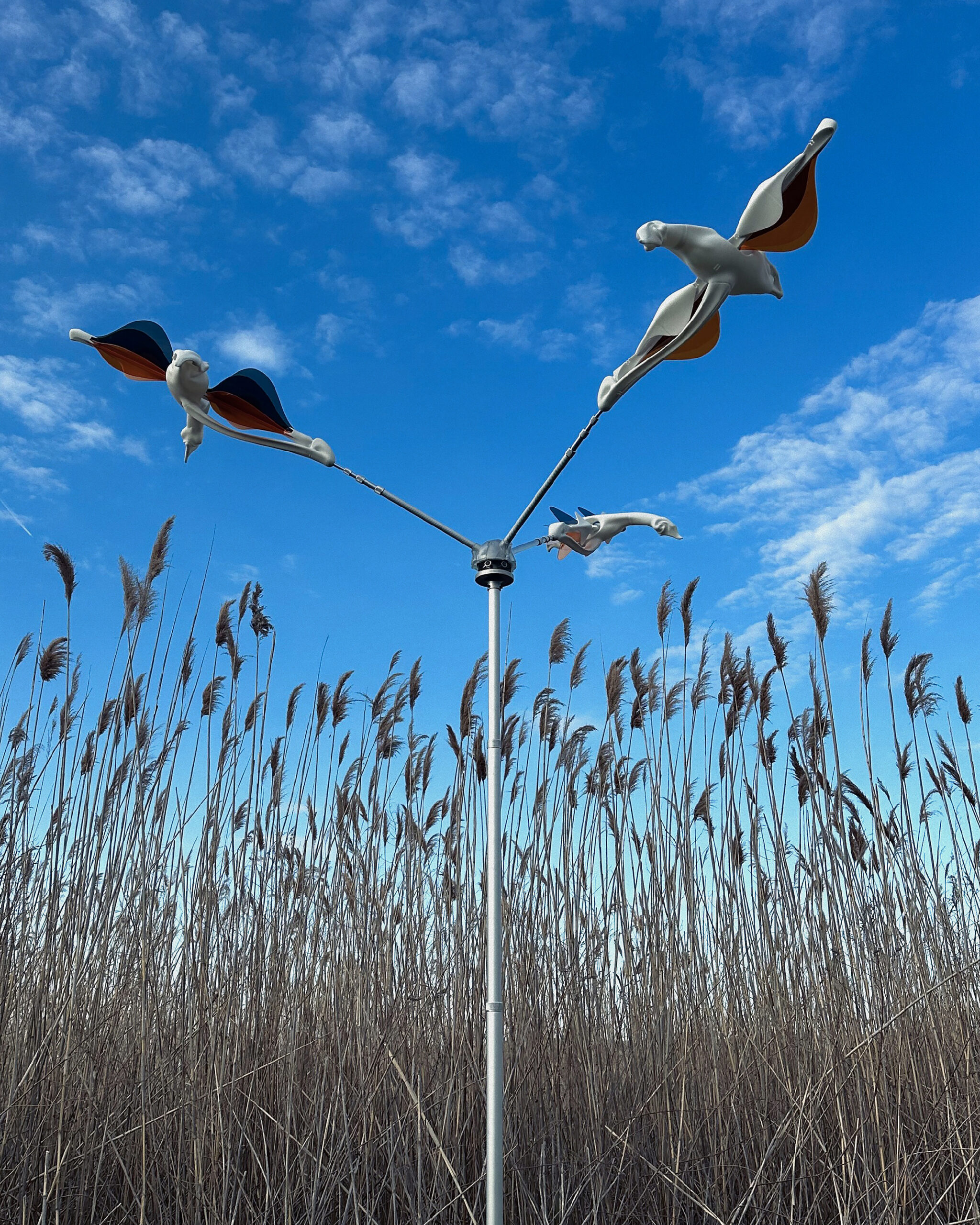 A decorative sculpture of three stylized birds in flight is mounted on a metal pole, set against a backdrop of tall, golden grasses. The sky above is blue with scattered clouds. The birds have striking colors, including orange and blue.