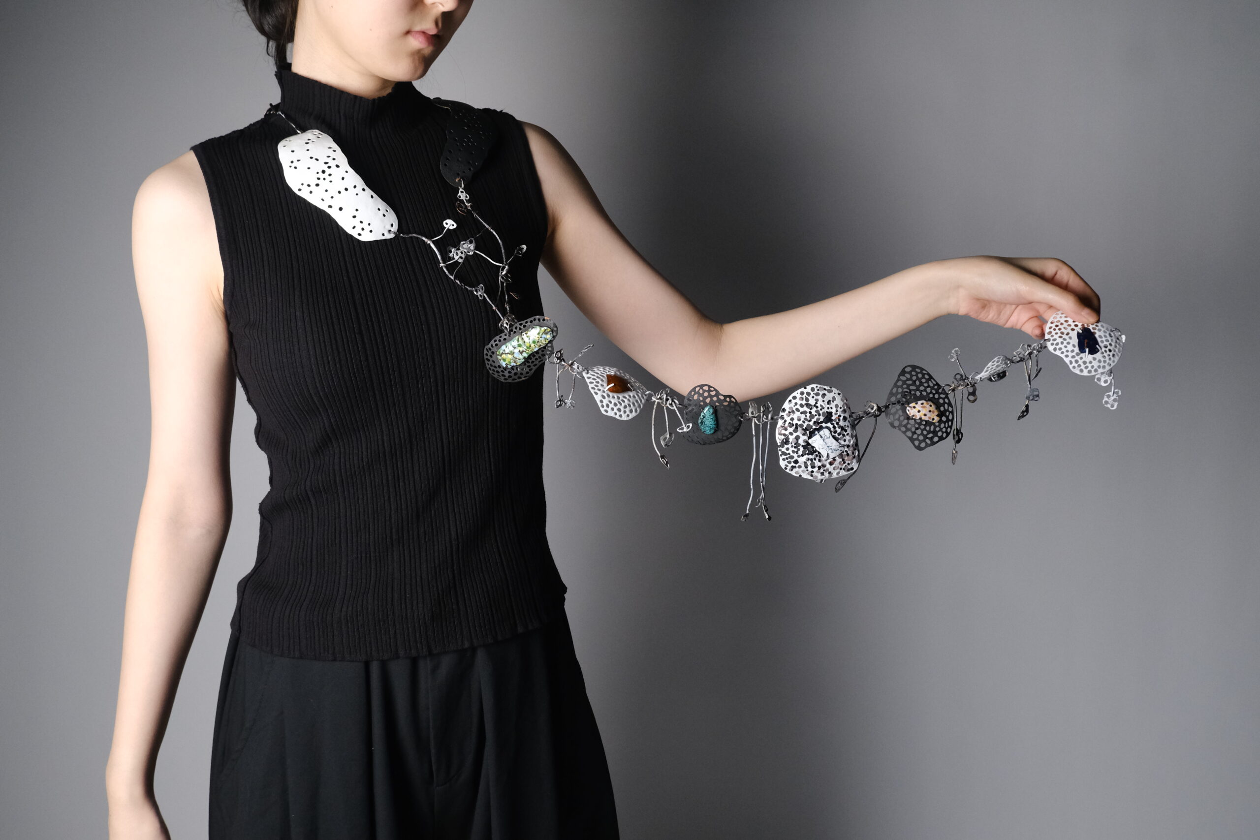 A person is wearing a sleeveless black top and is holding out an artistic necklace made of various textured and patterned elements resembling stones. The necklace features both solid and transparent pieces in shades of black, white, and silver, along with small decorative charms, all strung together in a creative design. The background is a gradient gray, emphasizing the necklace.