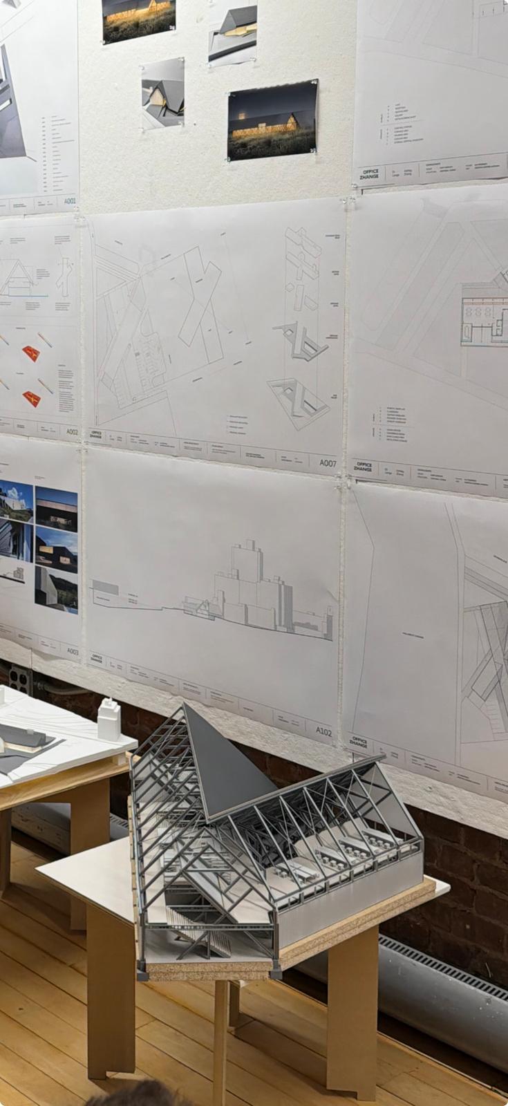 A detailed architectural model of a tall, narrow building with a textured exterior resembling vertical slats. The model is displayed on a black base against a backdrop of various architectural drawings and models. The design showcases an open interior with multiple levels and staircases, emphasizing modern design elements.