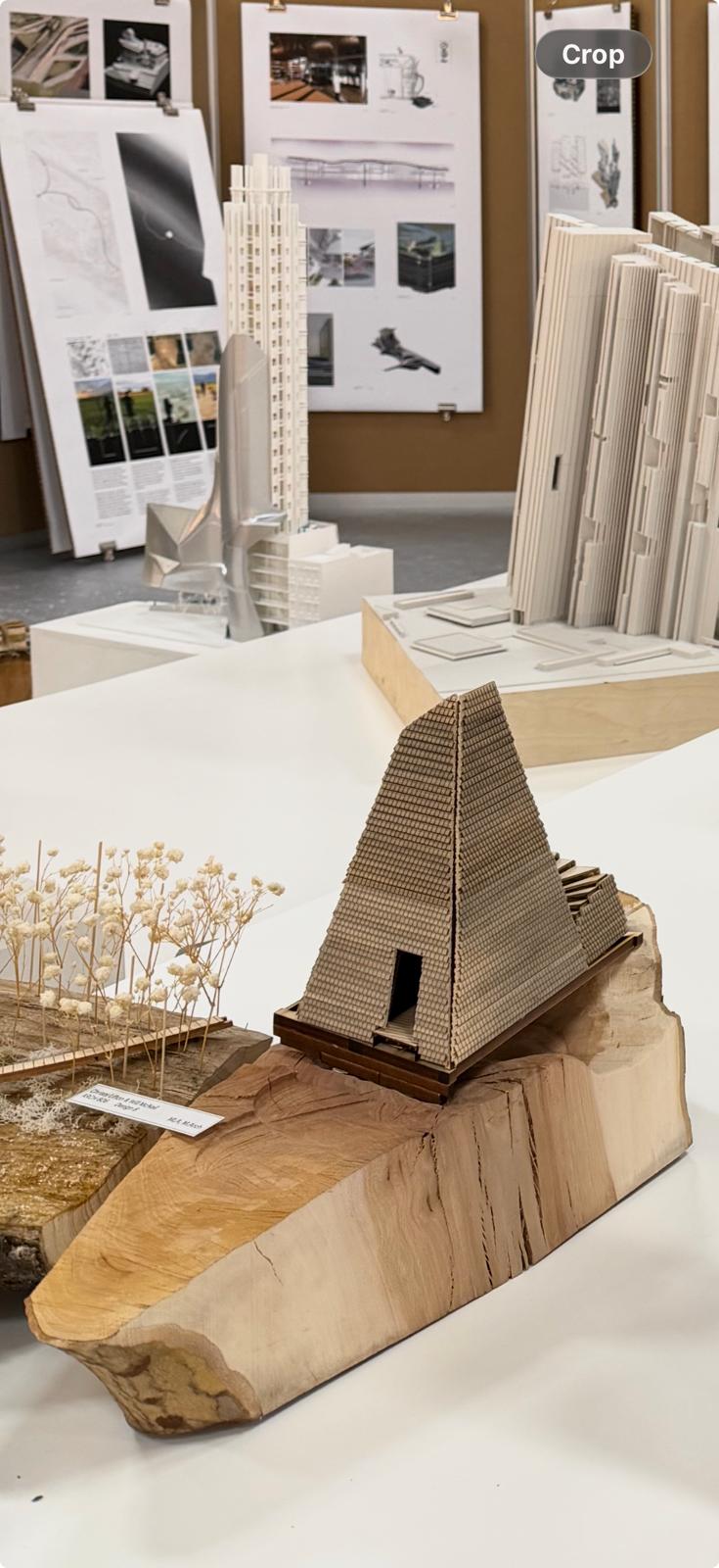 A detailed architectural model of a pyramid-shaped structure made of layered materials sits on a wooden base. To the left, a small arrangement of dried flowers is displayed on a piece of textured wood. In the background, various architectural sketches and models are pinned on a board, showcasing different designs and structures.