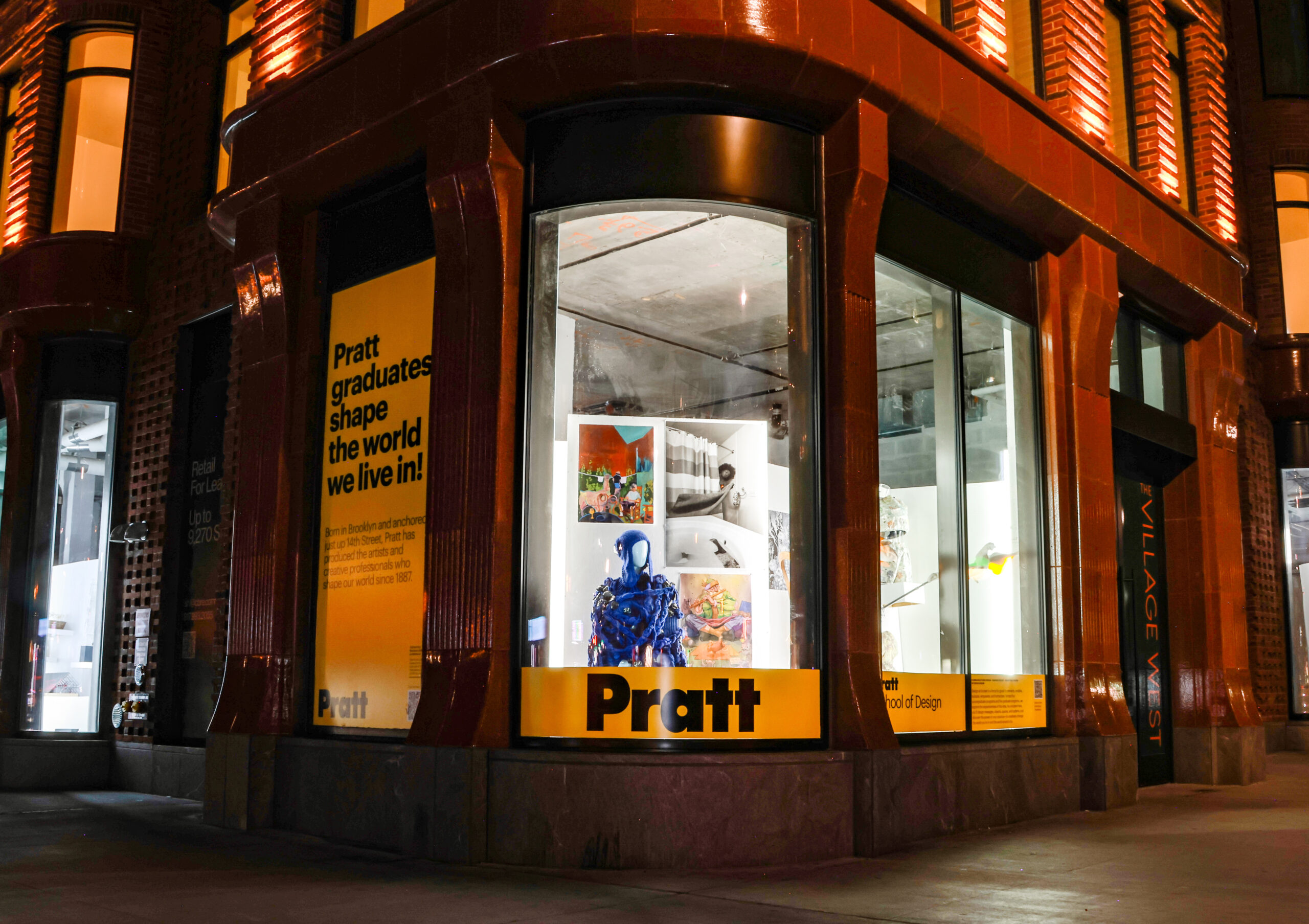 A brightly lit storefront displays a window exhibit for Pratt Institute at night. The window features colorful artwork and a prominent blue sculpture. A large yellow sign states, "Pratt graduates shape the world we live in!" with additional information about the institute. The exterior has dark brown architectural details, and a nearby sign indicates "The Village West."