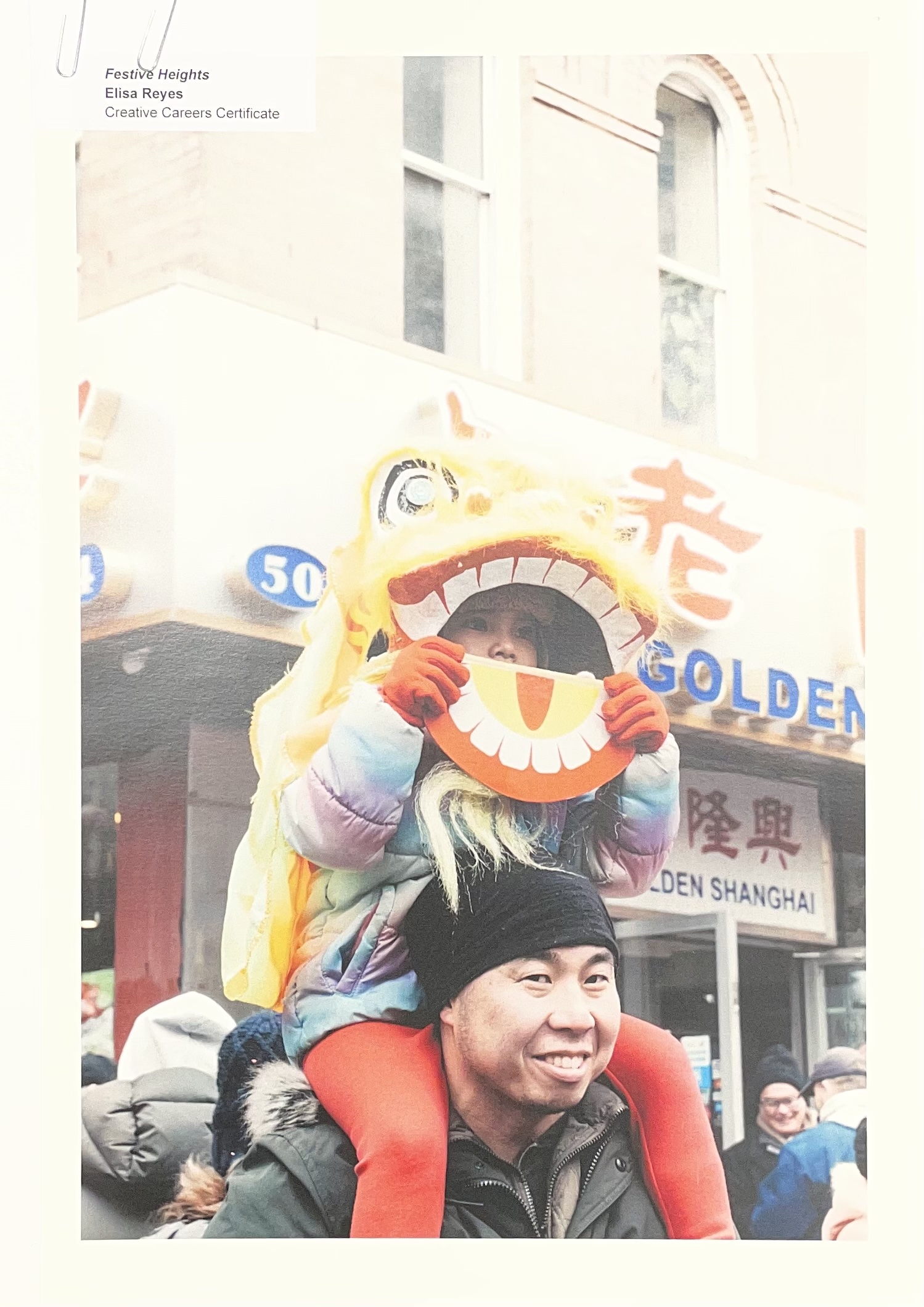 A young child wearing a colorful lion mask sits on the shoulders of an adult man, both smiling. The child holds a large, orange, smiling mask. In the background, there are signs for a restaurant, partially visible, and a crowd of people. The scene appears to be festive, likely during a parade or celebration.