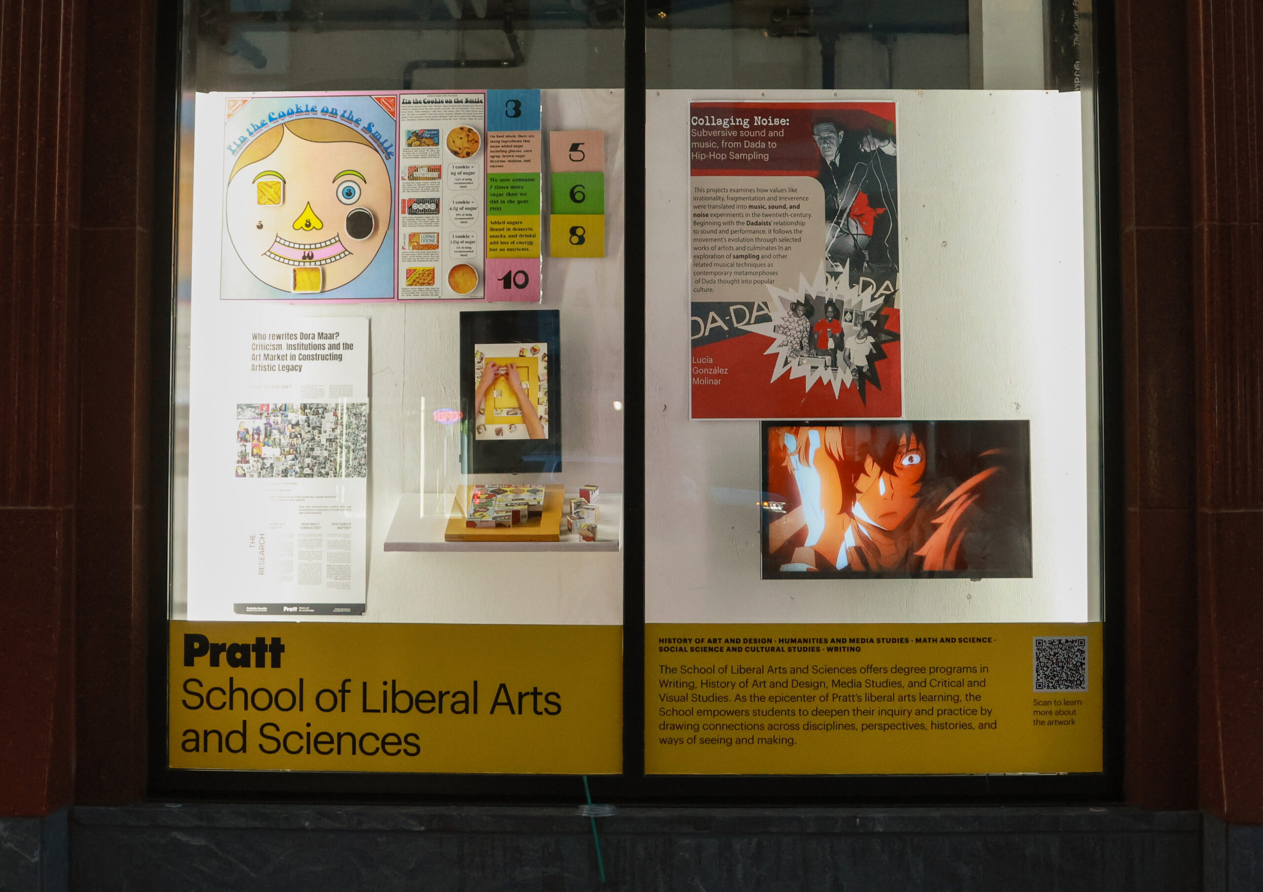 A window display featuring artwork from the Pratt School of Art. On the left, there is a striking painting of a muscular figure with a fragmented face against a colorful backdrop. To the right, various photographs showcase diverse subjects, including a black-and-white image of two figures in a field, and a close-up of a person holding an artistic accessory. The display includes a text panel about the school and a QR code for more information.