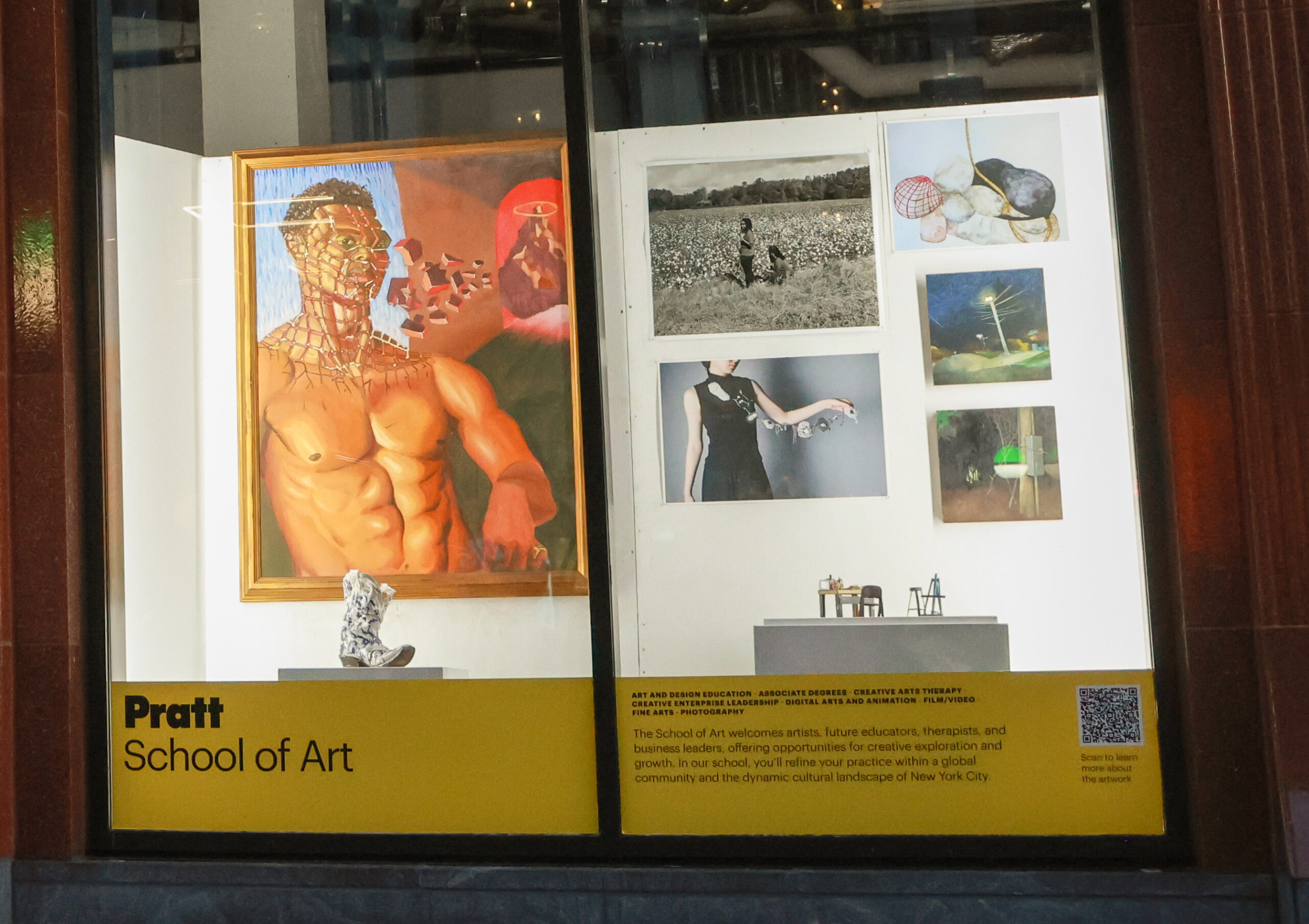 A window display features artwork from the Pratt School of Art. On the left, a large painting depicts a muscular figure with a fragmented head, surrounded by abstract shapes and a red hue. To the right, various photographs include a black-and-white image of two people in a field, a figure in a dark dress holding objects, and nature scenes. At the bottom, informational text about the school and a QR code for more details are visible.