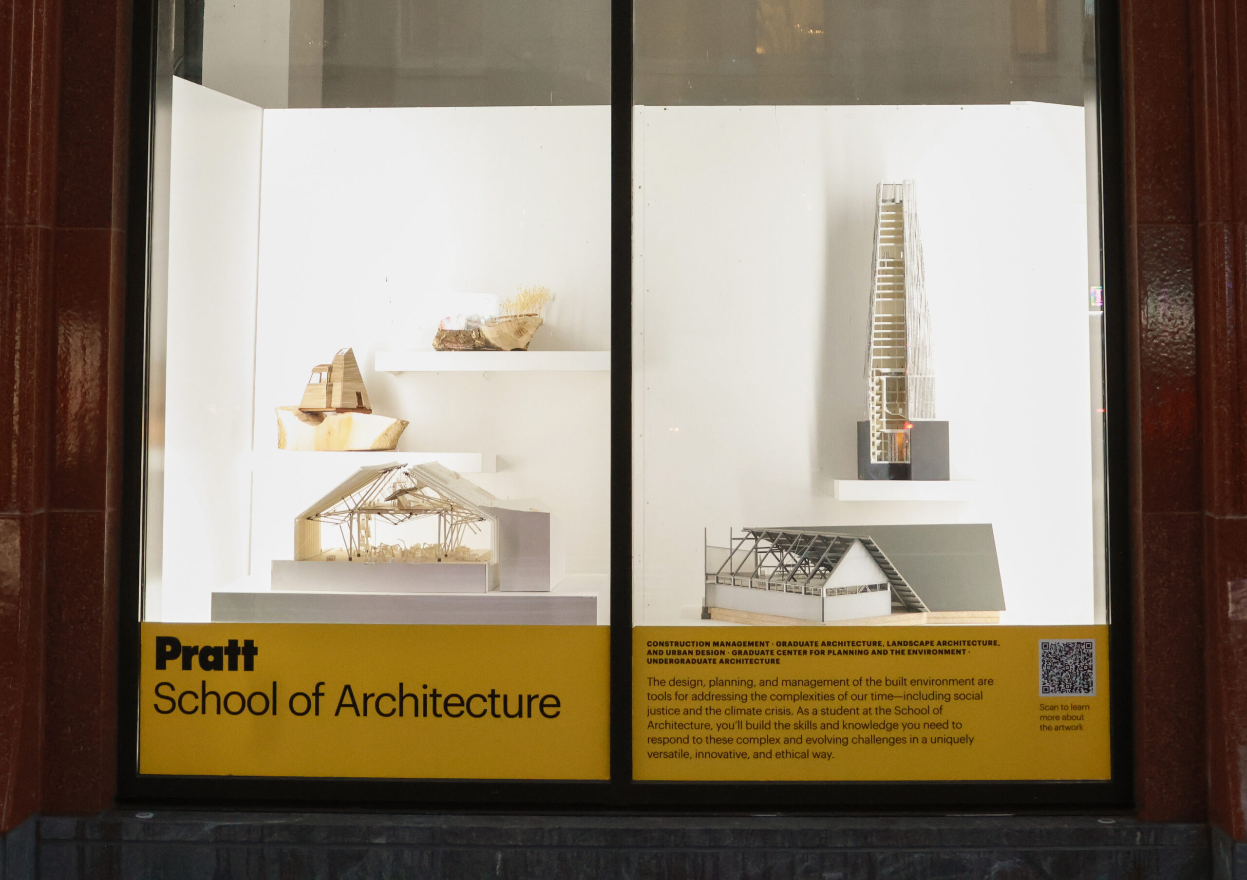 A storefront display for the Pratt School of Art featuring various artworks. On the left, a large painting of a muscular figure appears to break apart, while on the right, several photographs and artworks are hung, including a black-and-white image of people in a field and a color image of a figure with an unusual accessory. Below, a sign provides information about the school’s programs and encourages scanning a QR code for more details.