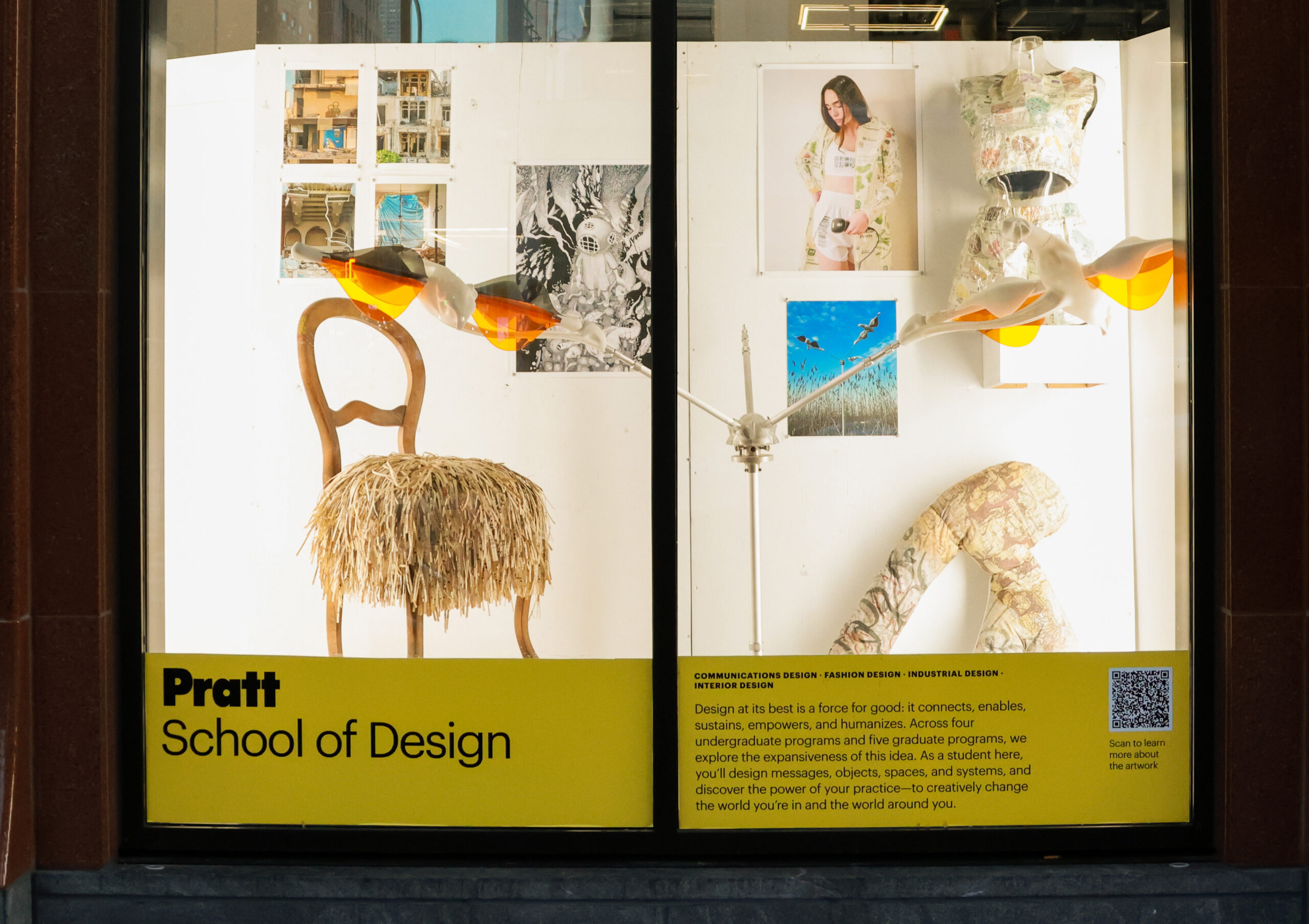 The image shows a window display for the Pratt School of Art, featuring a large painting of a muscular figure with broken pieces coming off, alongside various photographs and artworks on the adjacent wall. A sculptural piece is placed in front, and the display includes informational text about the school at the bottom, highlighting its programs and focus on art education.