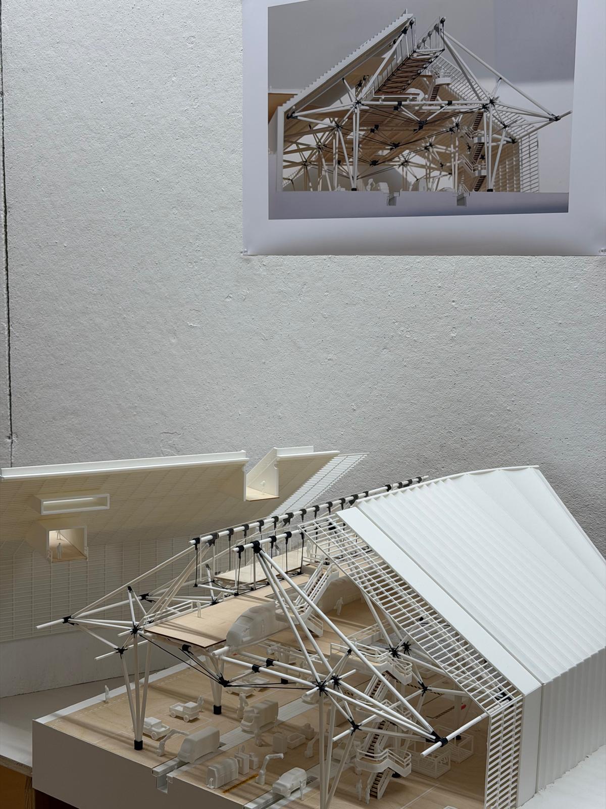 A detailed architectural model is displayed in the foreground, featuring a complex structure with white materials and visible interior elements. Above the model, a poster shows a rendered image of the same structure, highlighting its design and features. The background is a simple textured wall, emphasizing the model and poster.
