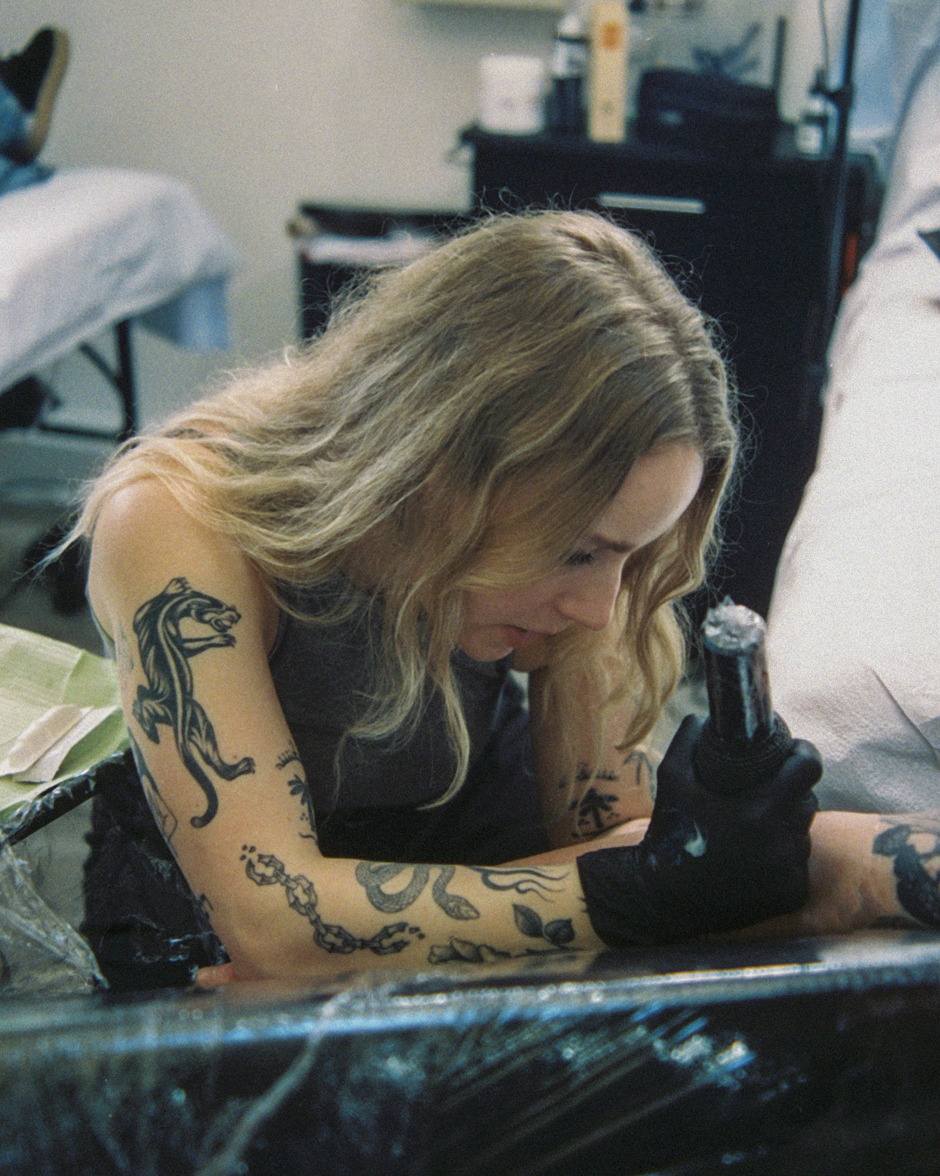 A tattoo artist with long, wavy hair and multiple tattoos on their arms is focused on tattooing. They are holding a tattoo machine in one hand and leaning over a client's arm on a tattoo bed, which is covered with plastic wrap. The background includes some tattoo equipment and a chair.