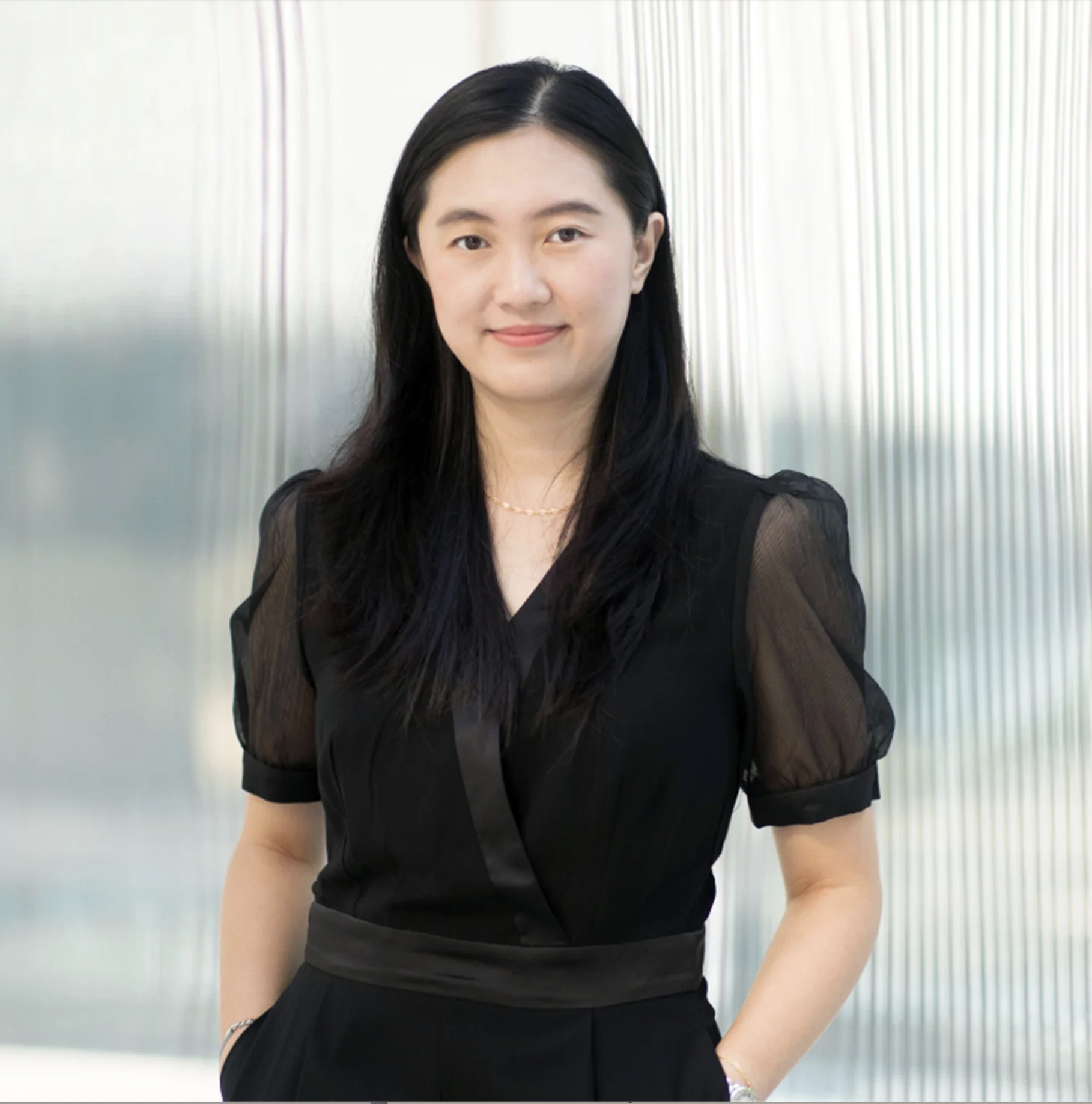 A woman stands confidently in front of a textured, glass-like background. She has long black hair and wears a black dress with puffed sleeves and a waist belt. Her hands are positioned in her pockets, and she has a subtle smile. The lighting is bright, enhancing the professional atmosphere of the setting.