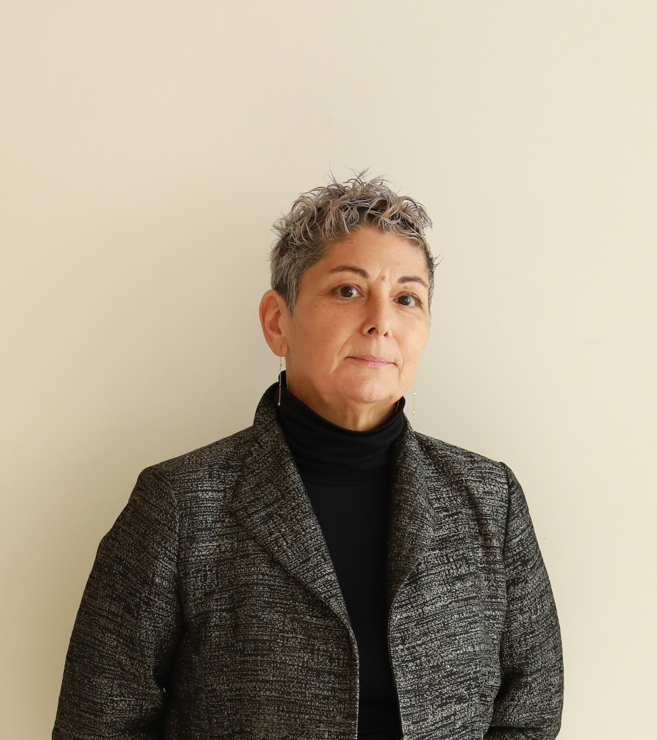 A woman with short, silver curly hair stands against a plain light-colored wall. She is wearing a black turtleneck and a textured dark blazer. The expression on her face is serious, and she appears confident and composed.