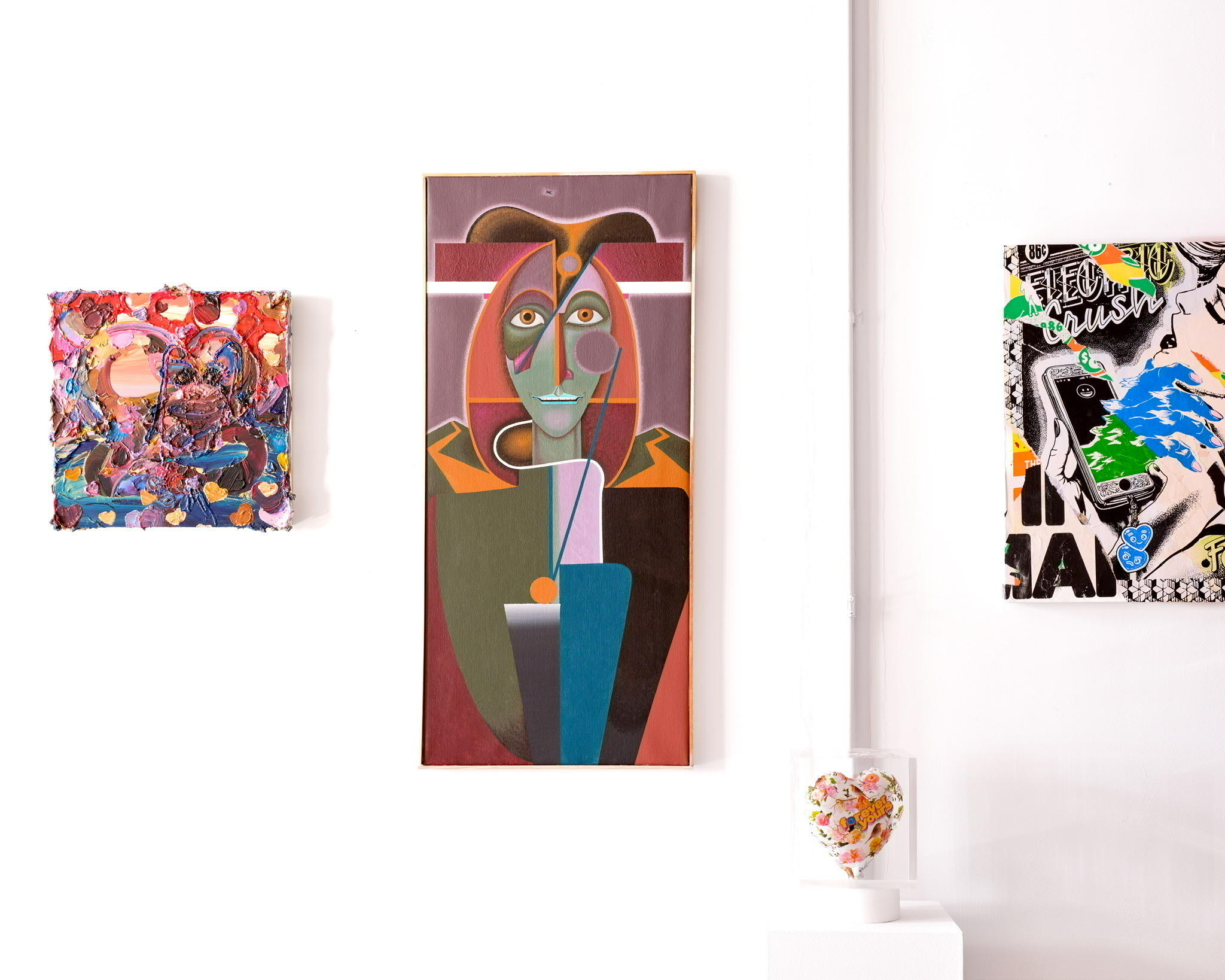 Three artworks are displayed on a white wall. On the left, a small textured painting with vibrant colors, patterns, and a heart motif. The central piece is a larger artwork depicting a stylized face with angular features, muted colors, and abstract elements. On the right, another artwork combines graphic design with bright blue and green elements, featuring a phone and text. A decorative heart-shaped object in a glass base is placed on a white pedestal in the foreground.