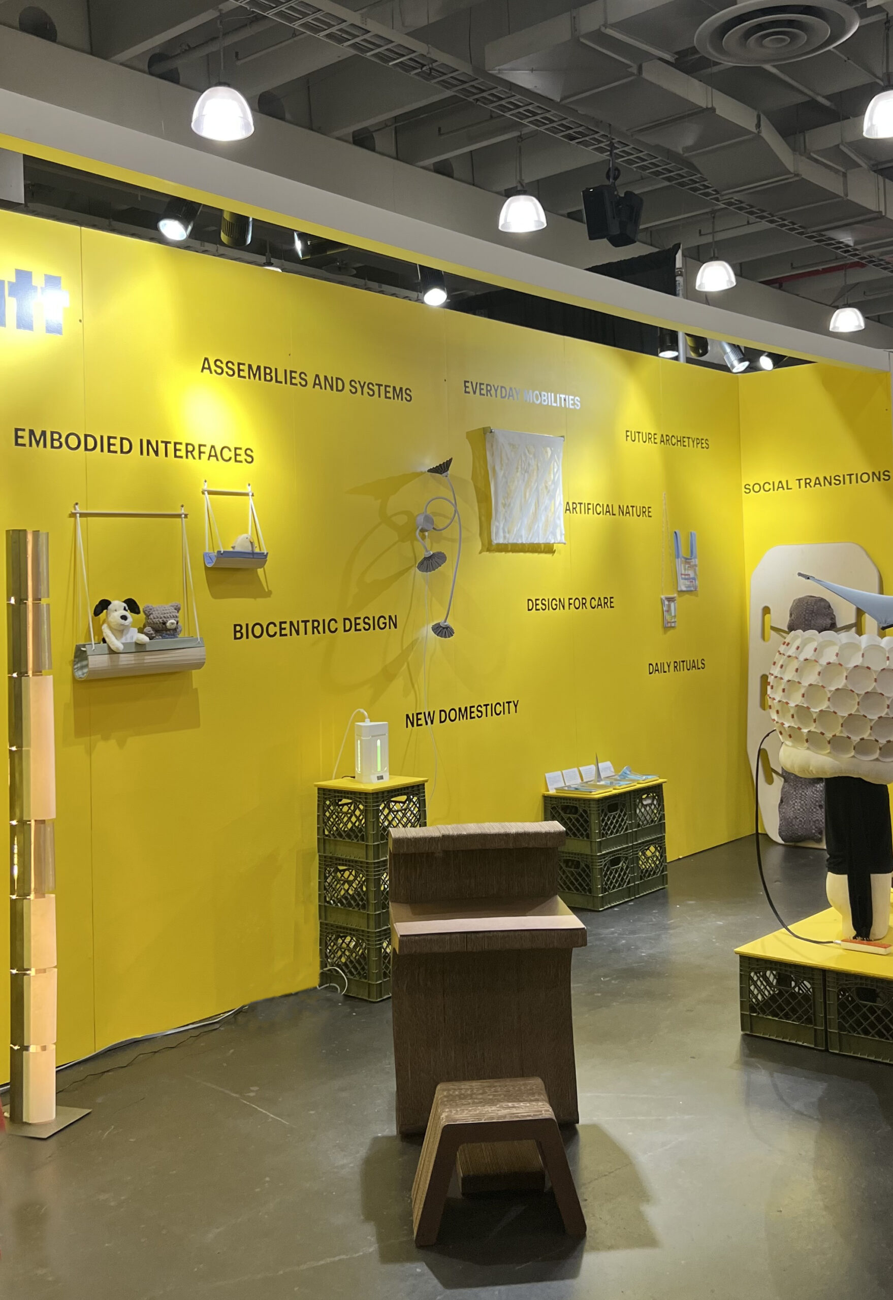 A vibrant yellow exhibition wall displays various themes in design, including "Assemblies and Systems," "Everyday Mobilities," and "Biocentric Design." Below the text, there are several design elements: a light fixture, decorative items, a small table, and a chair made from crates. Plush toys are visible on a shelf, contributing to the overall theme of domesticity and innovation in everyday objects. The space is well-lit, highlighting the contemporary design.