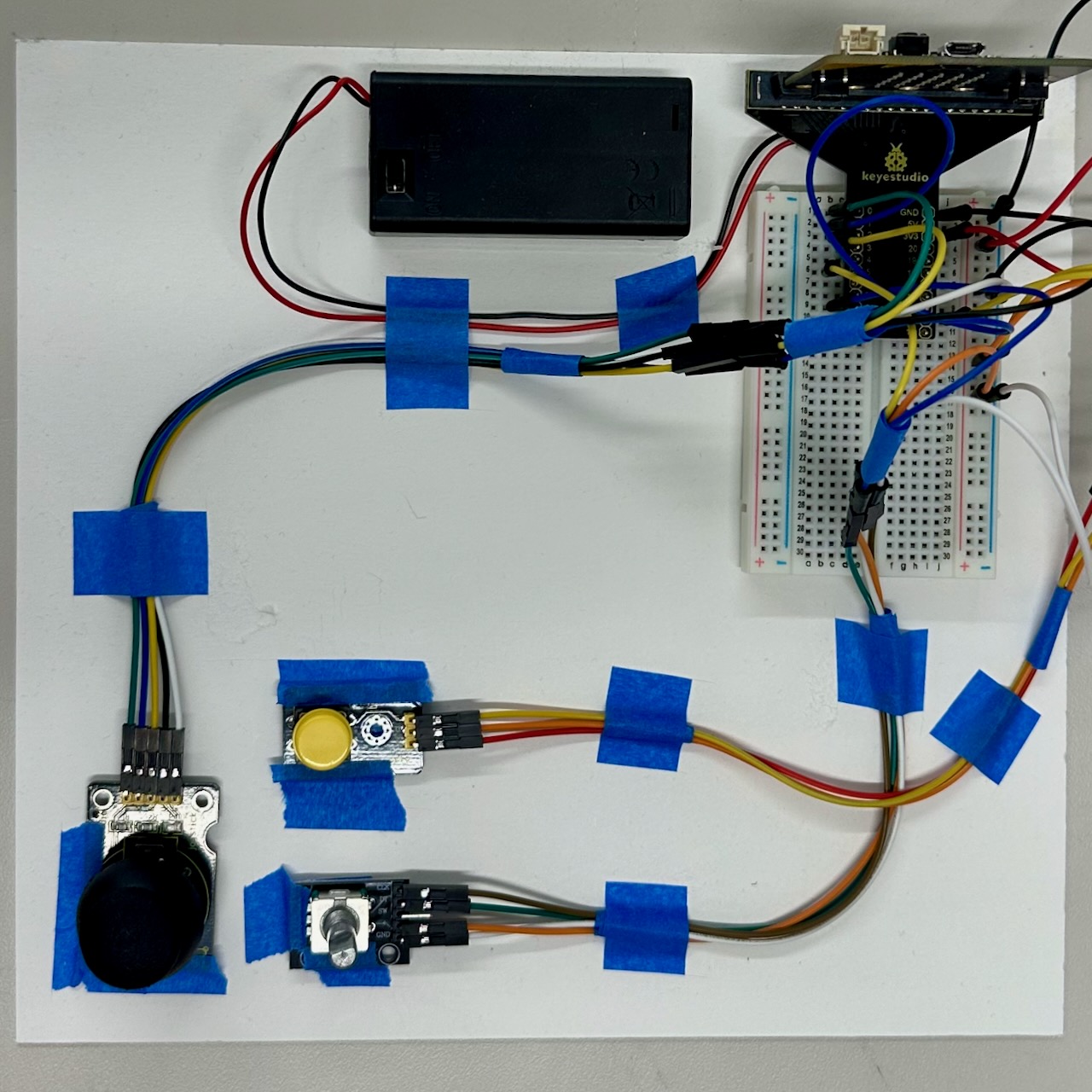 A flat setup featuring a breadboard connected to various electronic components. A battery pack is positioned at the top left, while multiple colored wires connect to a joystick, a potentiometer, and a button, all secured with blue tape. The breadboard displays several additional connections.