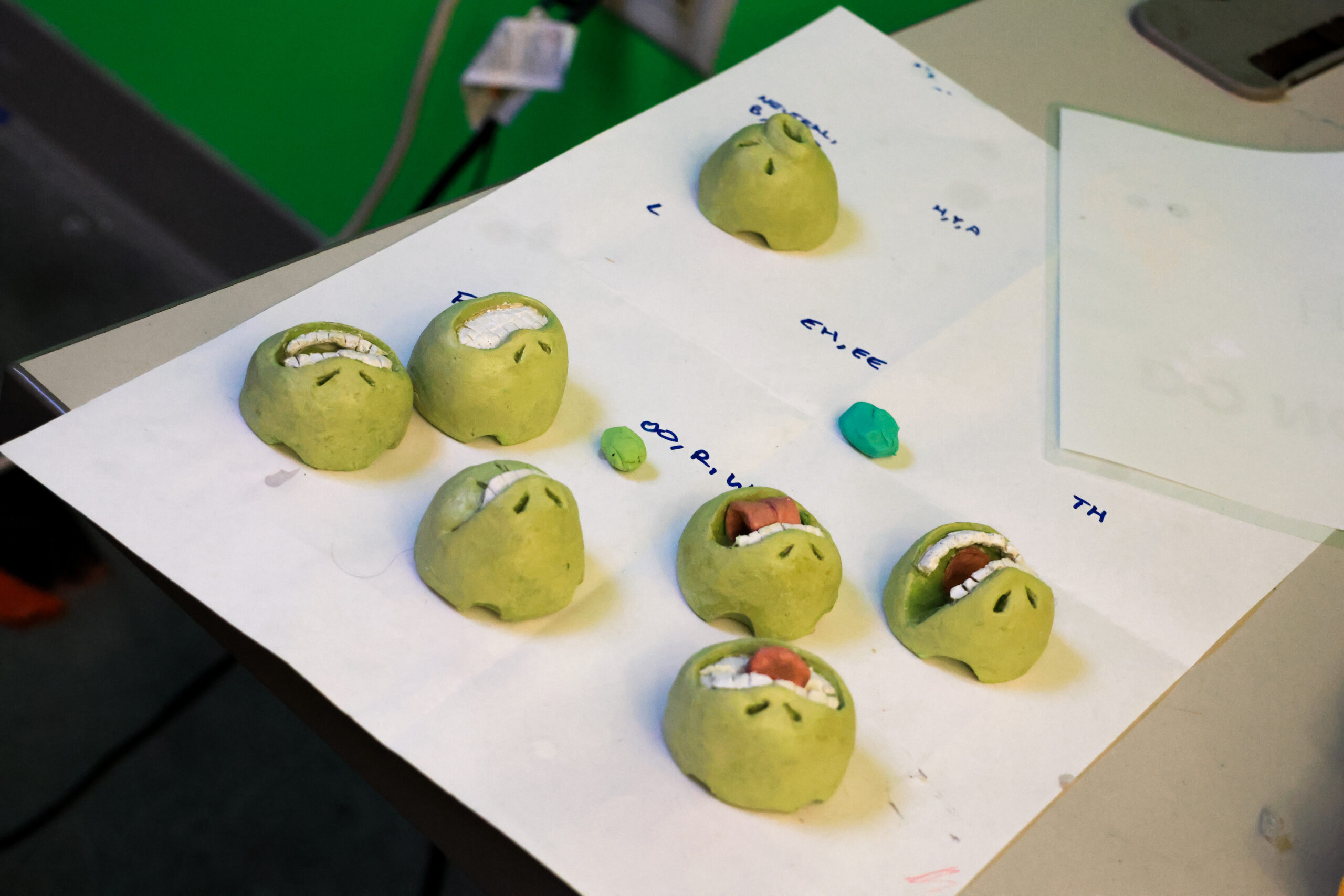 A collection of small green clay heads is arranged on a sheet of paper on a table. The heads have varying facial expressions, some showing teeth, while a few have colored detailing for the mouth. Nearby, there are small pieces of colored clay, including a blue piece. The background features a green wall and electrical outlets.