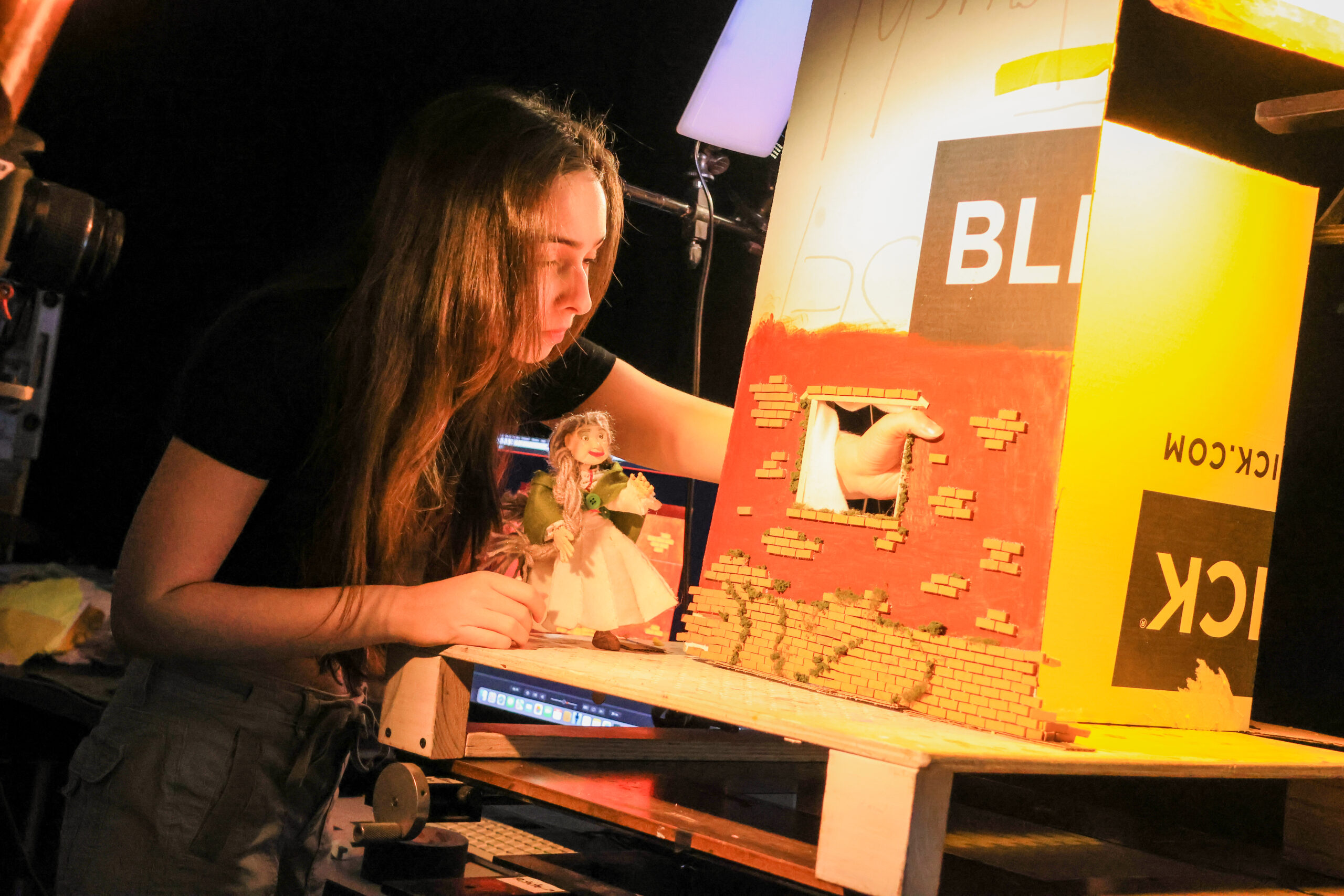 A young person is working on a diorama, focusing intently on a brick wall structure with a window. She holds a small doll in her other hand, positioned in front of a lit backdrop. A computer monitor is visible in the background, displaying tools or materials related to the project. The scene is well-lit, emphasizing her concentration and the details of the diorama.