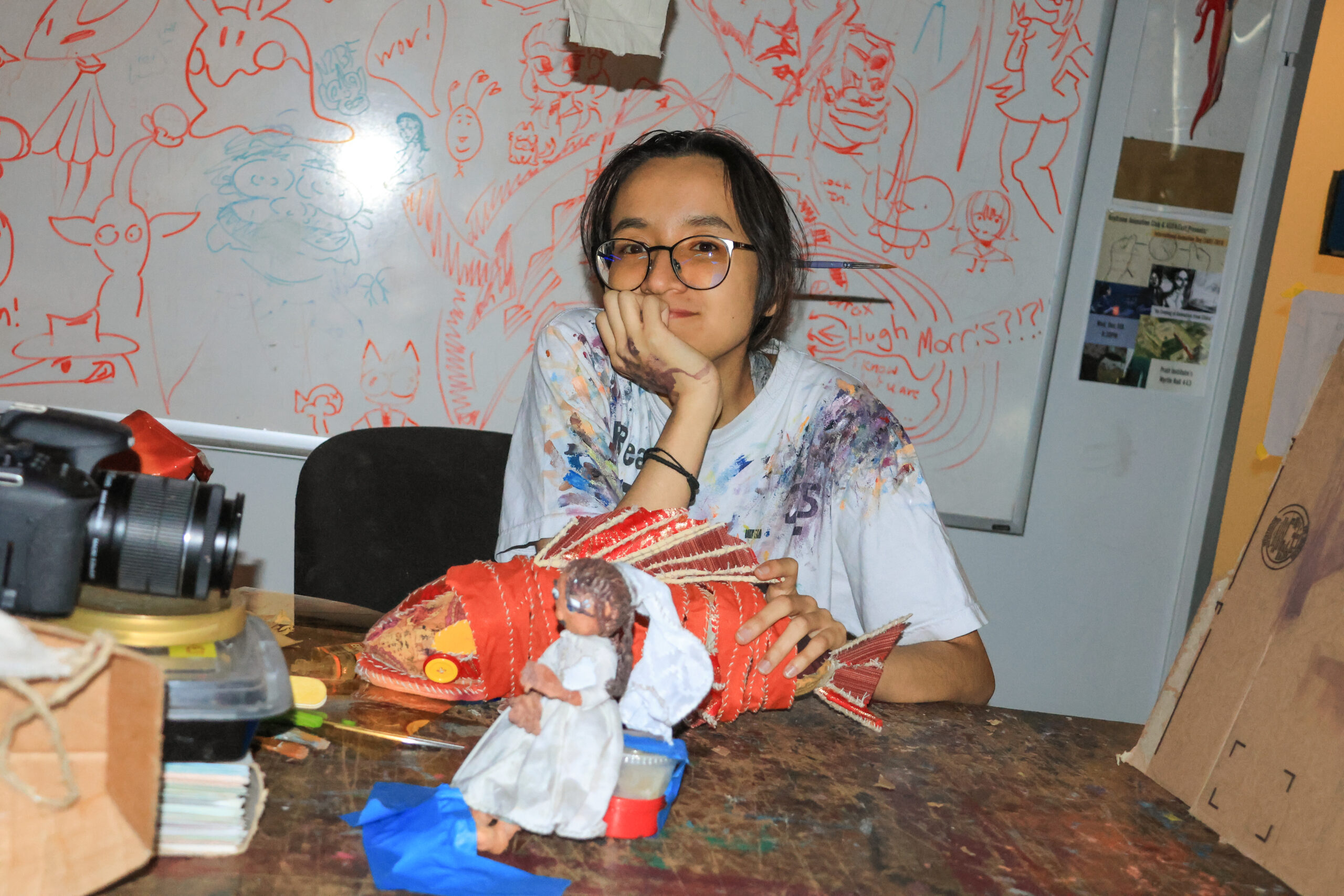 A person sits at a cluttered table, resting their chin on their hand, wearing a paint-splattered white t-shirt. In front of them is a large, handmade fish sculpture made of red material and a small doll dressed in white. A camera and various art supplies are visible on the table, with a colorful drawing board covered in sketches and doodles in the background.