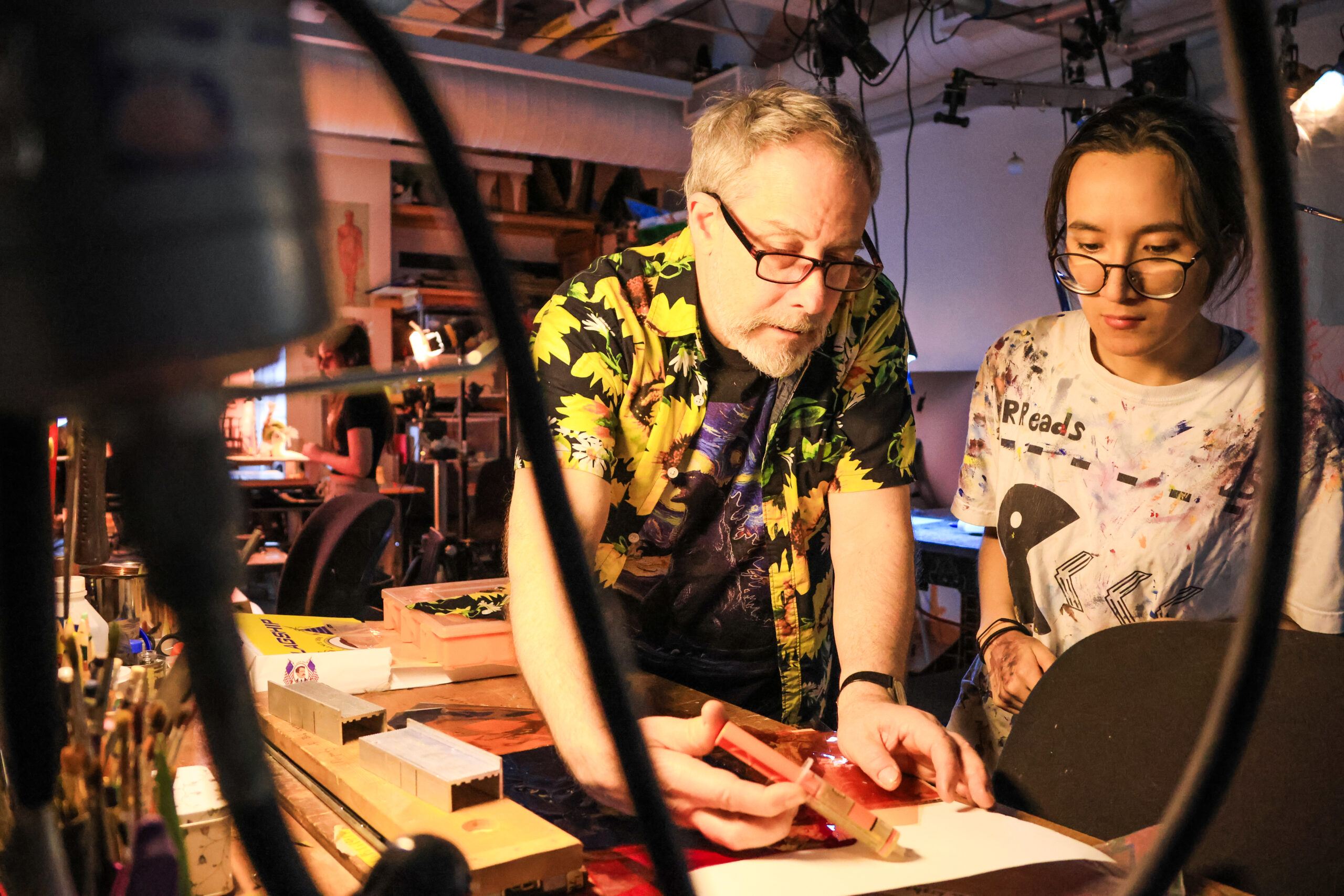 Two individuals are engaged in a creative activity at a workspace cluttered with art supplies. One person, wearing a colorful floral shirt, is using a red tool on paper, while the other, wearing a paint-splattered shirt, watches closely. The background features various art materials and equipment in a warmly lit studio environment.