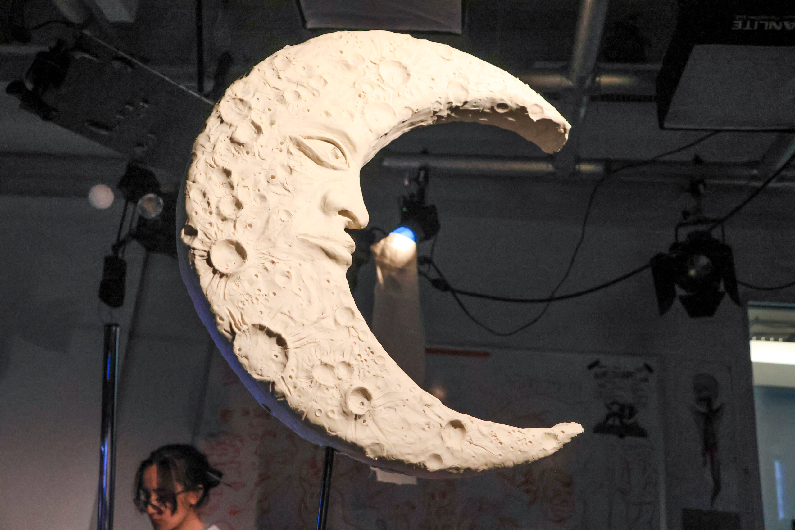 A sculpted crescent moon with a textured surface depicting craters and a face is showcased in a dimly lit studio. A person in the background, partially obscured, appears to be working on another piece. Several overhead lights illuminate the scene, casting shadows on the walls filled with sketches and drawings.